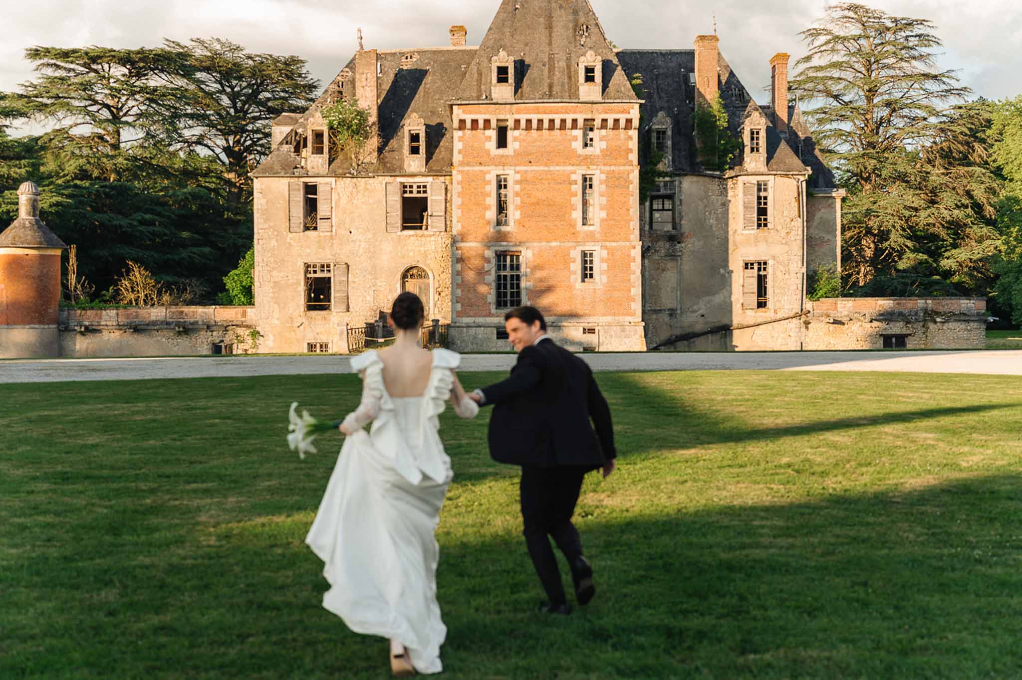 Bride and groom running hand-in-hand across lawn toward Renaissance chateau in golden-hour light