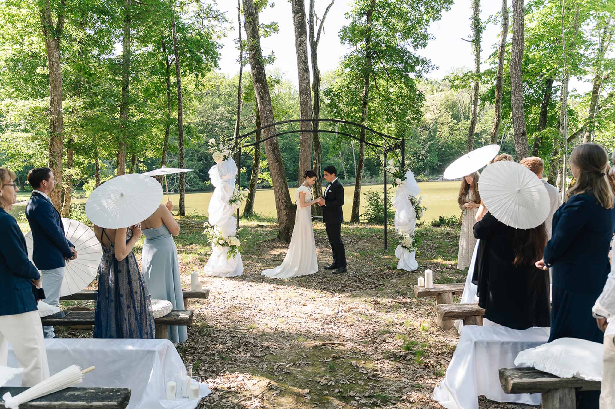 Couple exchanging vows under black metal arch with white draping in wooded clearing with candle-lit bench rows