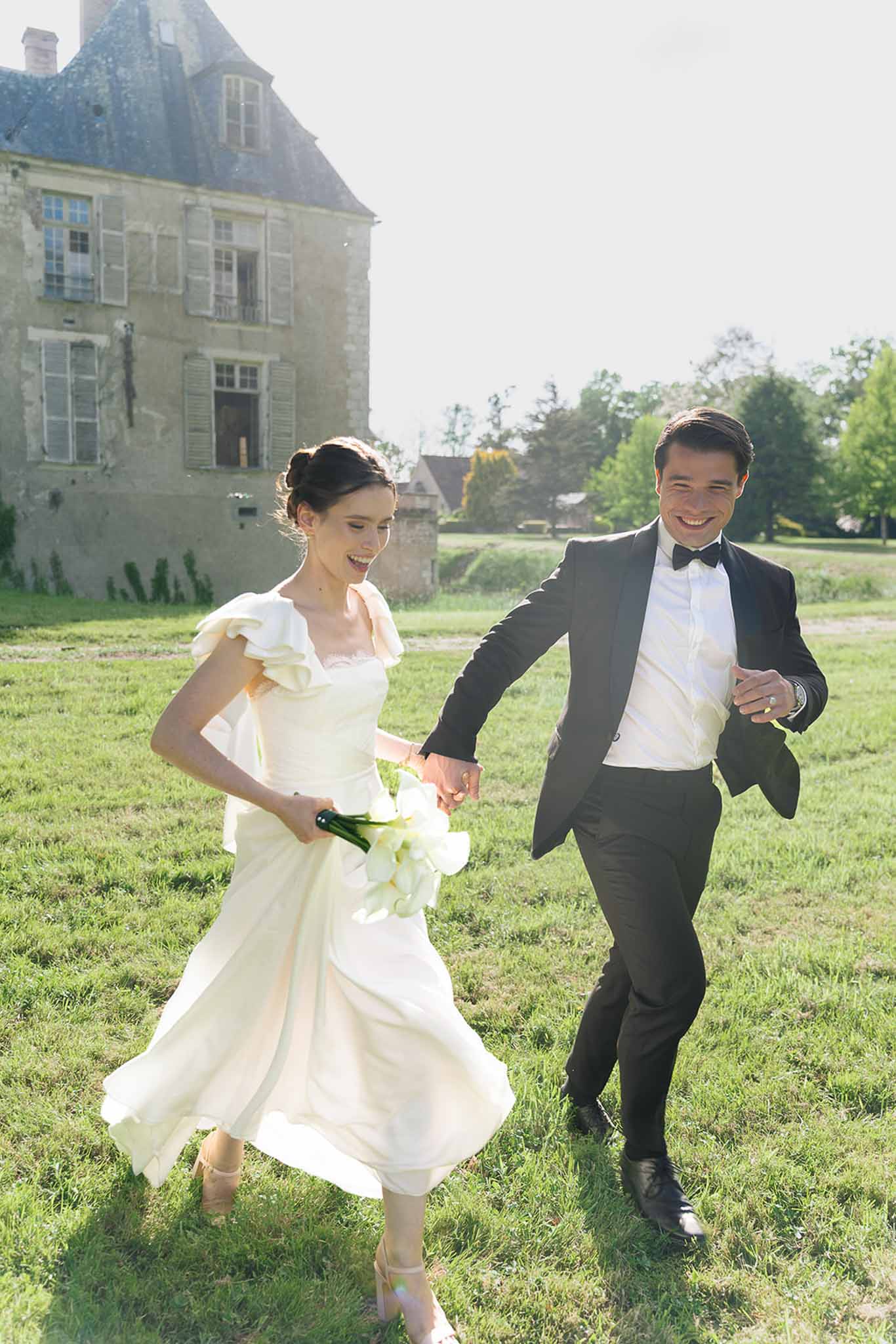 Bride and groom running hand-in-hand across chateau lawn, bride in ivory ruffled midi dress with calla lily bouquet