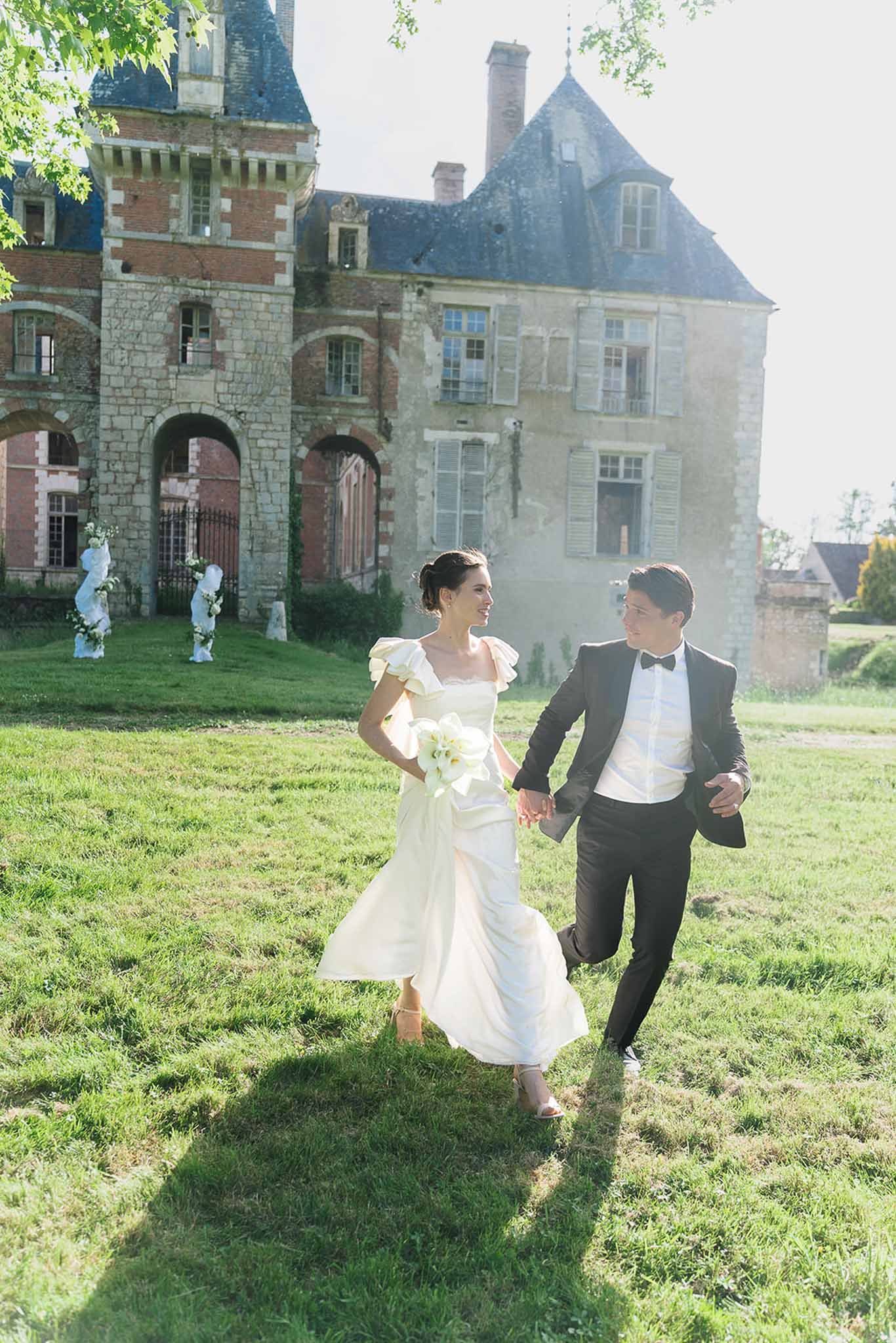 Bride and groom walking and laughing on lawn with white calla lily bouquet and chateau facade behind