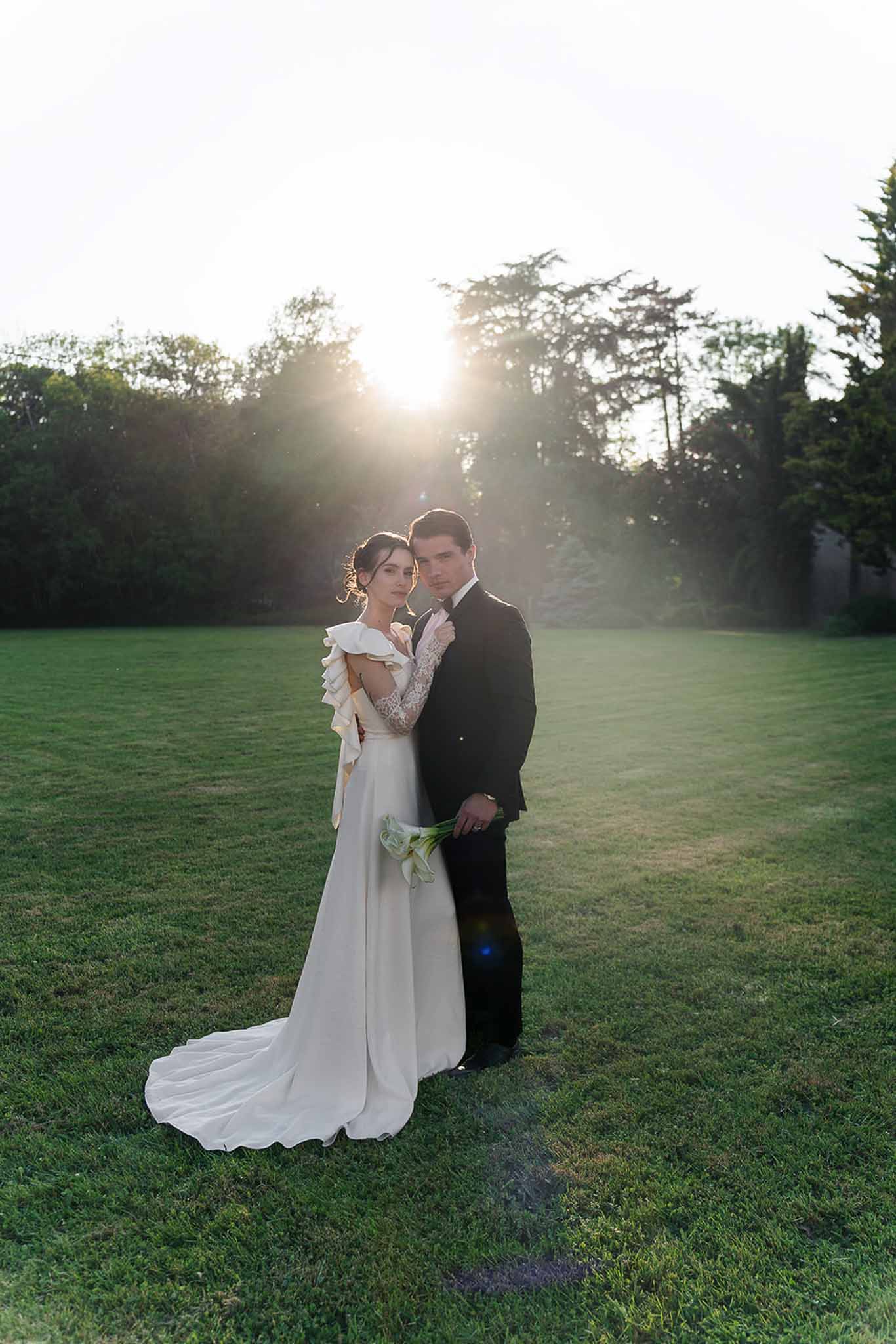 Couple with calla lily bouquet and ruffled-back gown poses at golden hour with sun flare on lawn