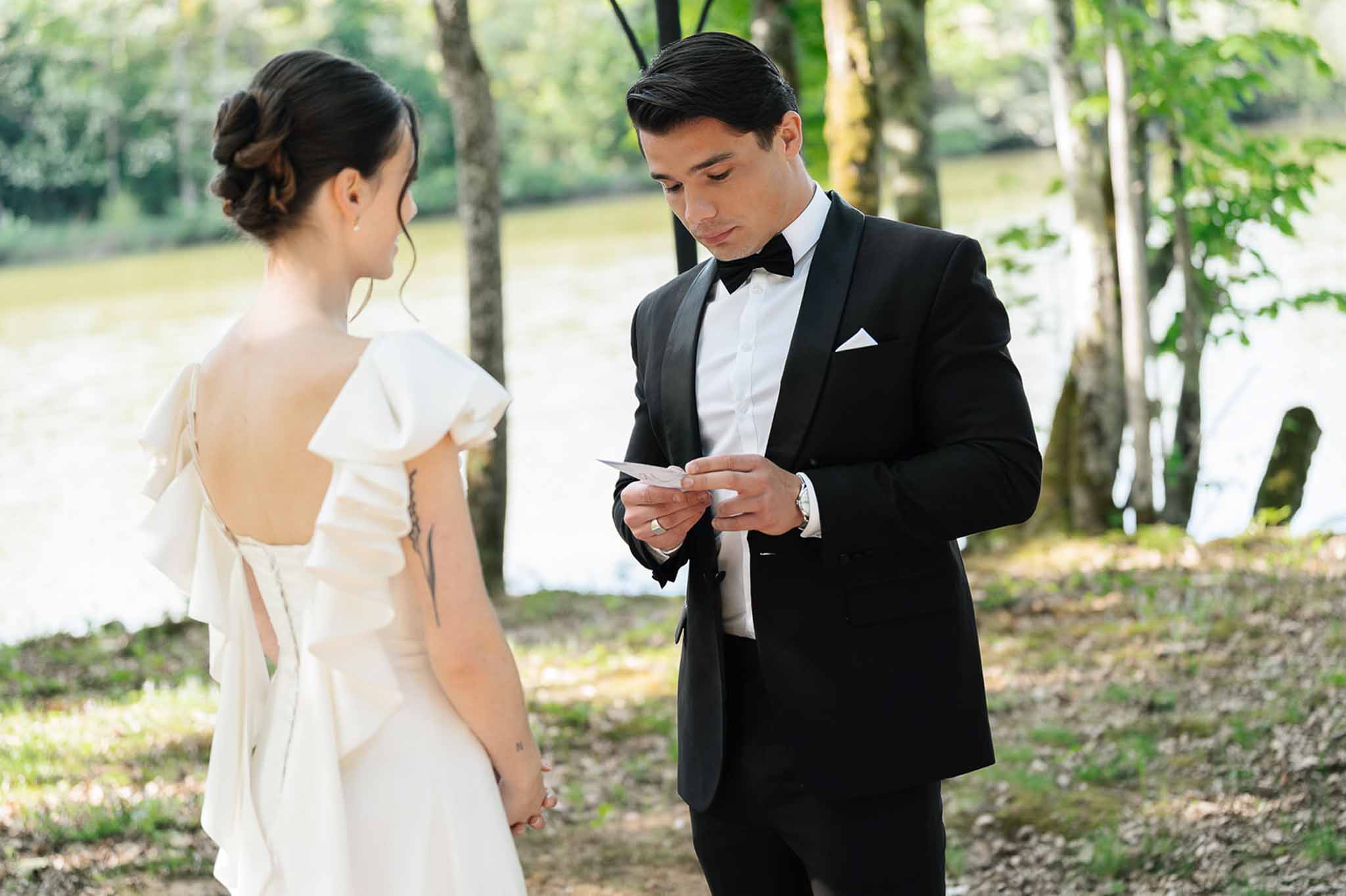 Couple exchanging private vows beside lake, groom reading card in tuxedo, bride in flutter-sleeve gown