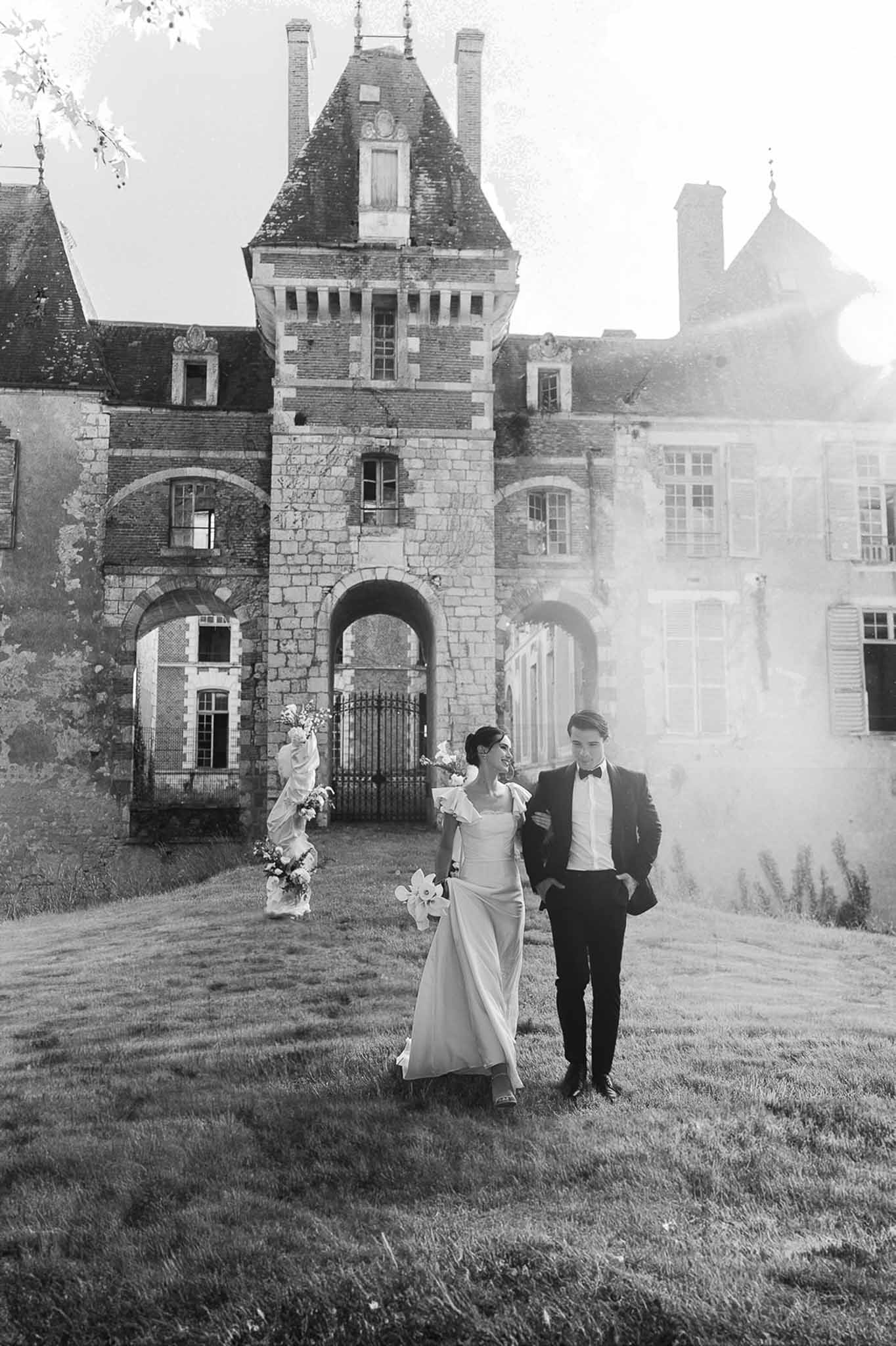 Black and white couple walking before turreted chateau bride in column gown with ruffle shoulders and bouquet