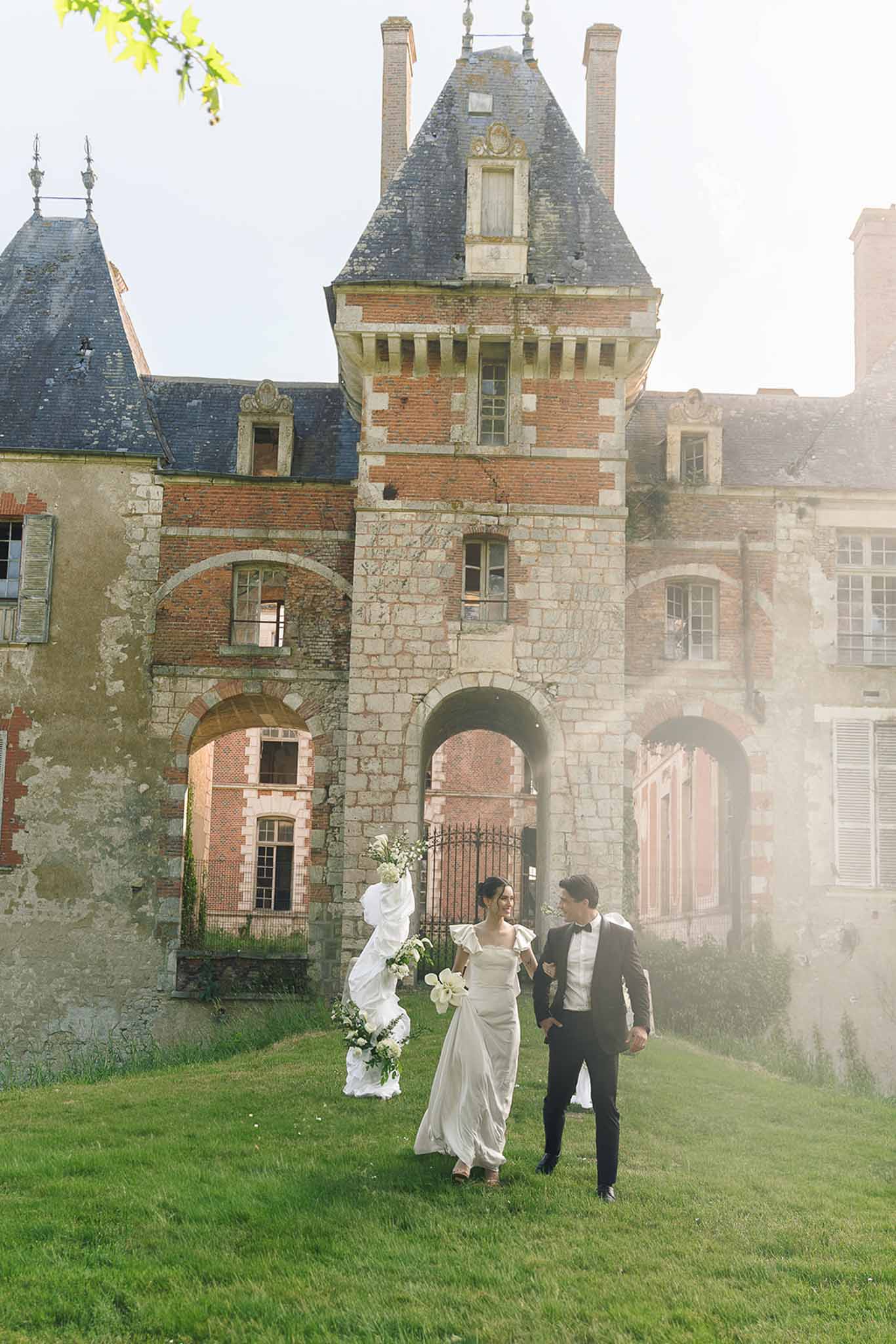 Couple walking across lawn with magnolia bouquet and billowing train before red brick chateau with slate turrets