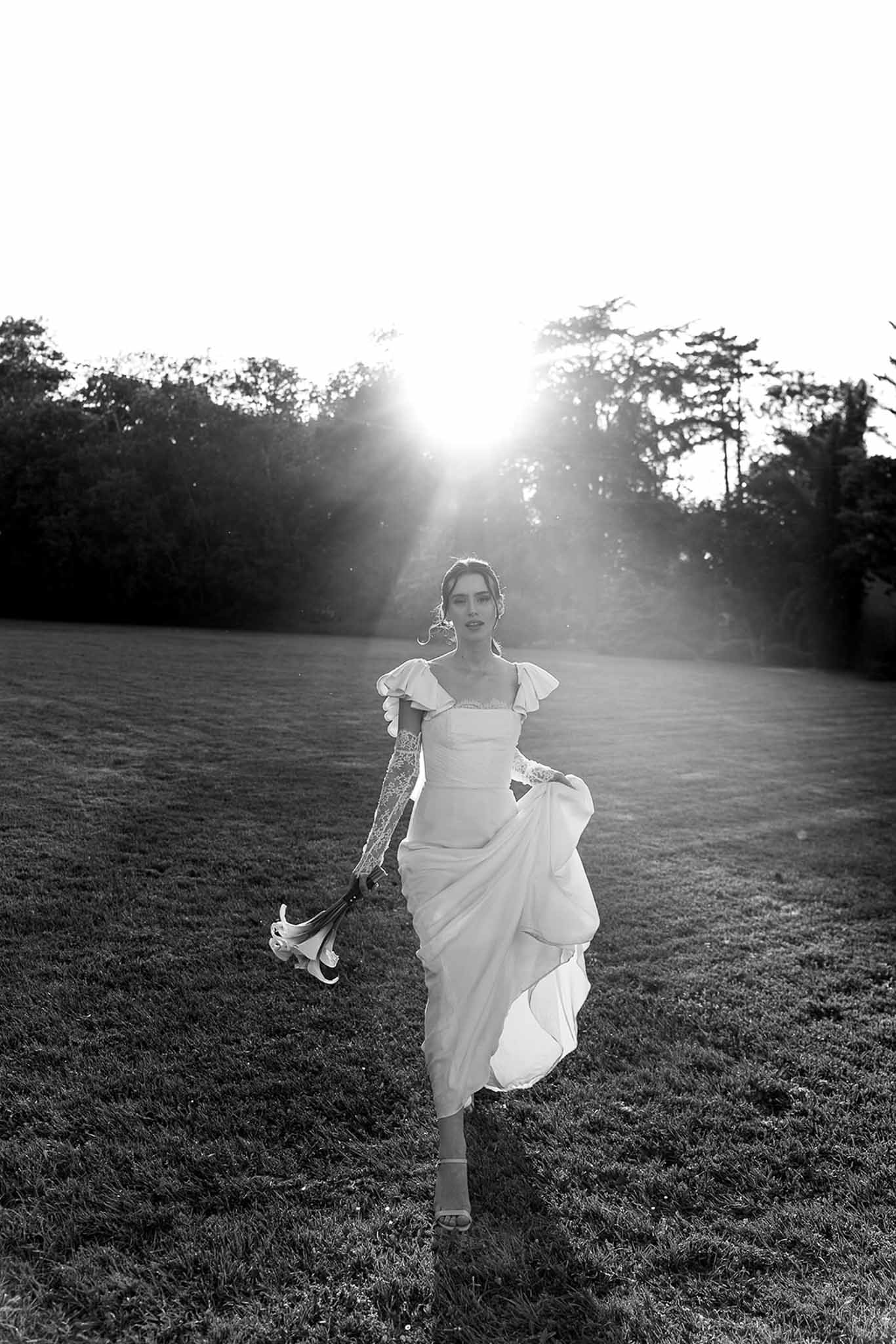 Black and white portrait of bride walking across lawn in midi-length gown with lace gloves backlit by low sun