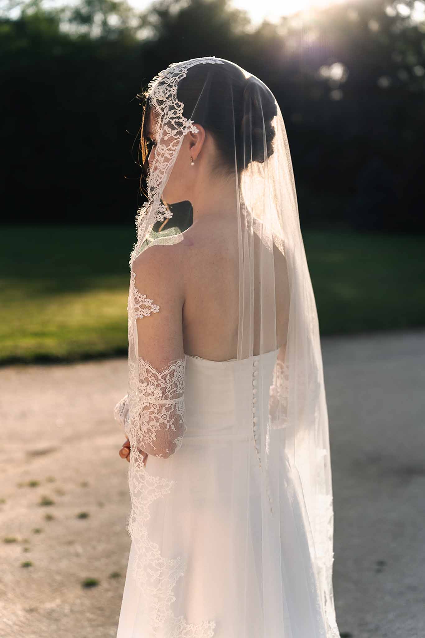 Backlit bridal portrait showing low-back ivory gown with lace sleeves and chapel-length lace-trimmed veil