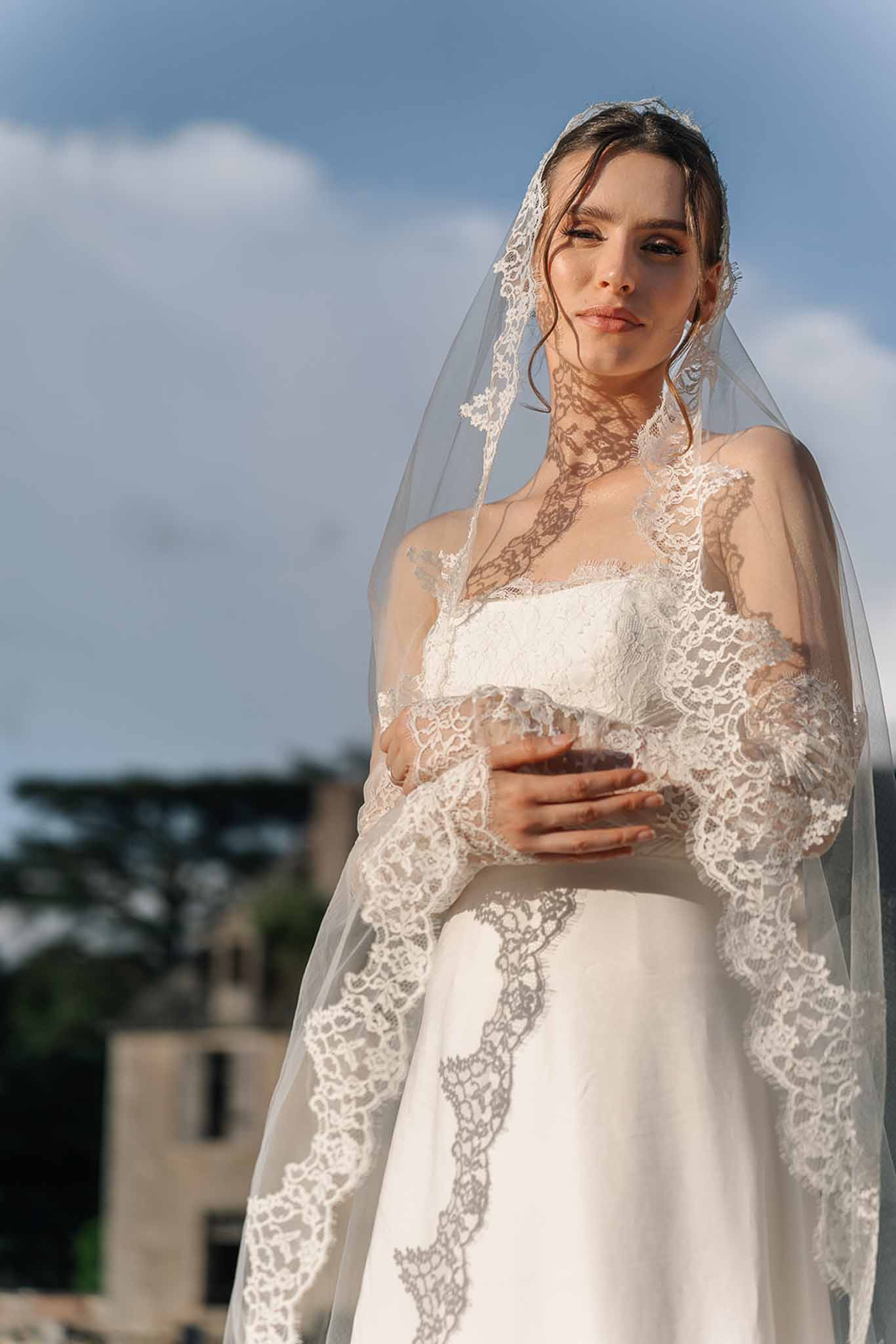 Bride in Chantilly lace-edged cathedral veil holding red wine glass against open sky