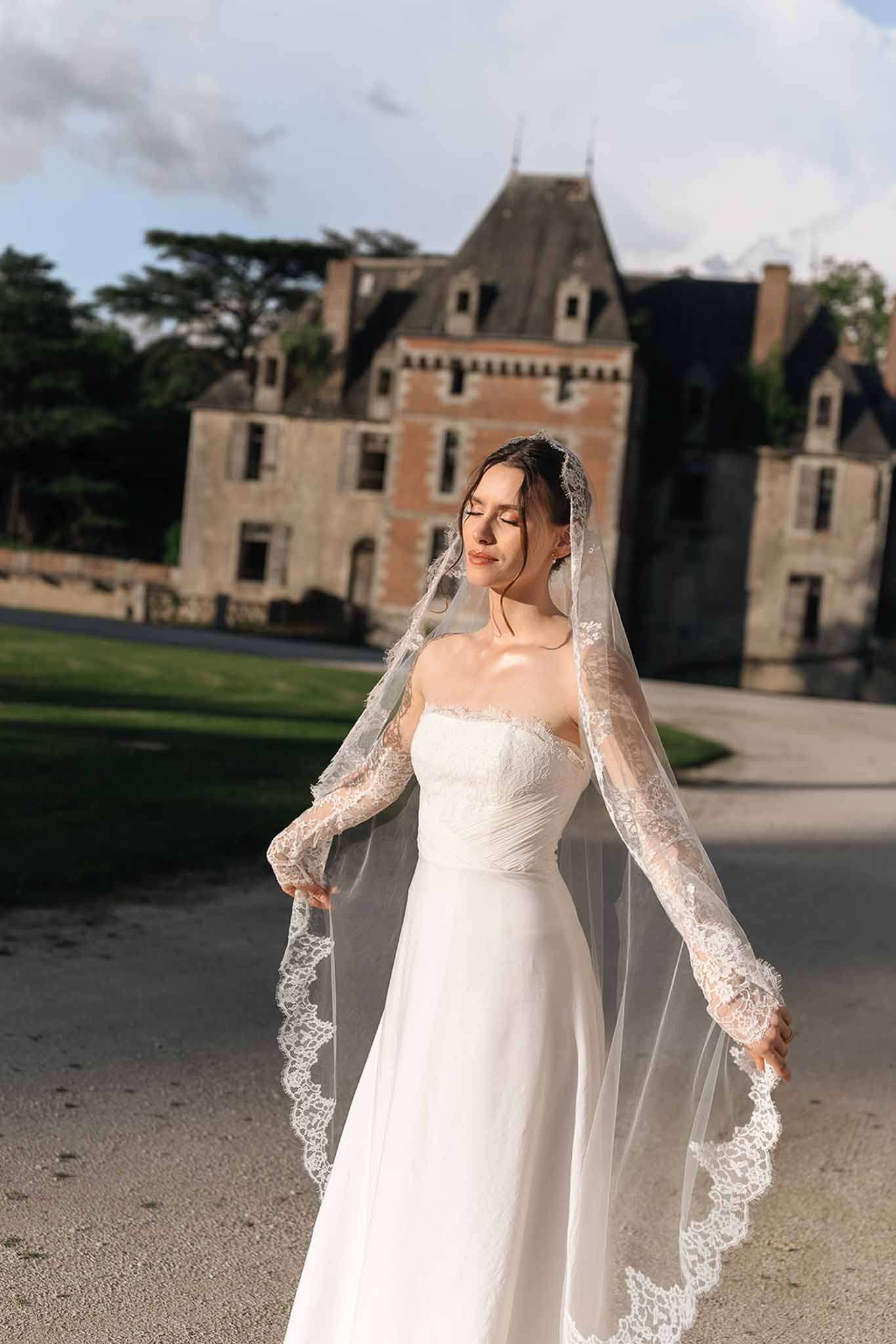 Bride holding cathedral-length lace veil on chateau gravel driveway in warm afternoon light
