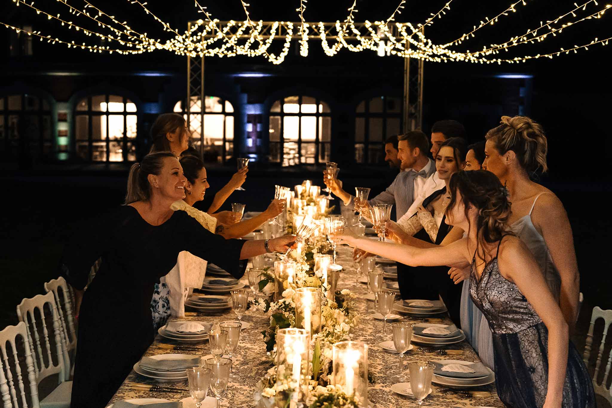 Ten guests toasting at candlelit table with greenery runner under dense fairy light canopy at night