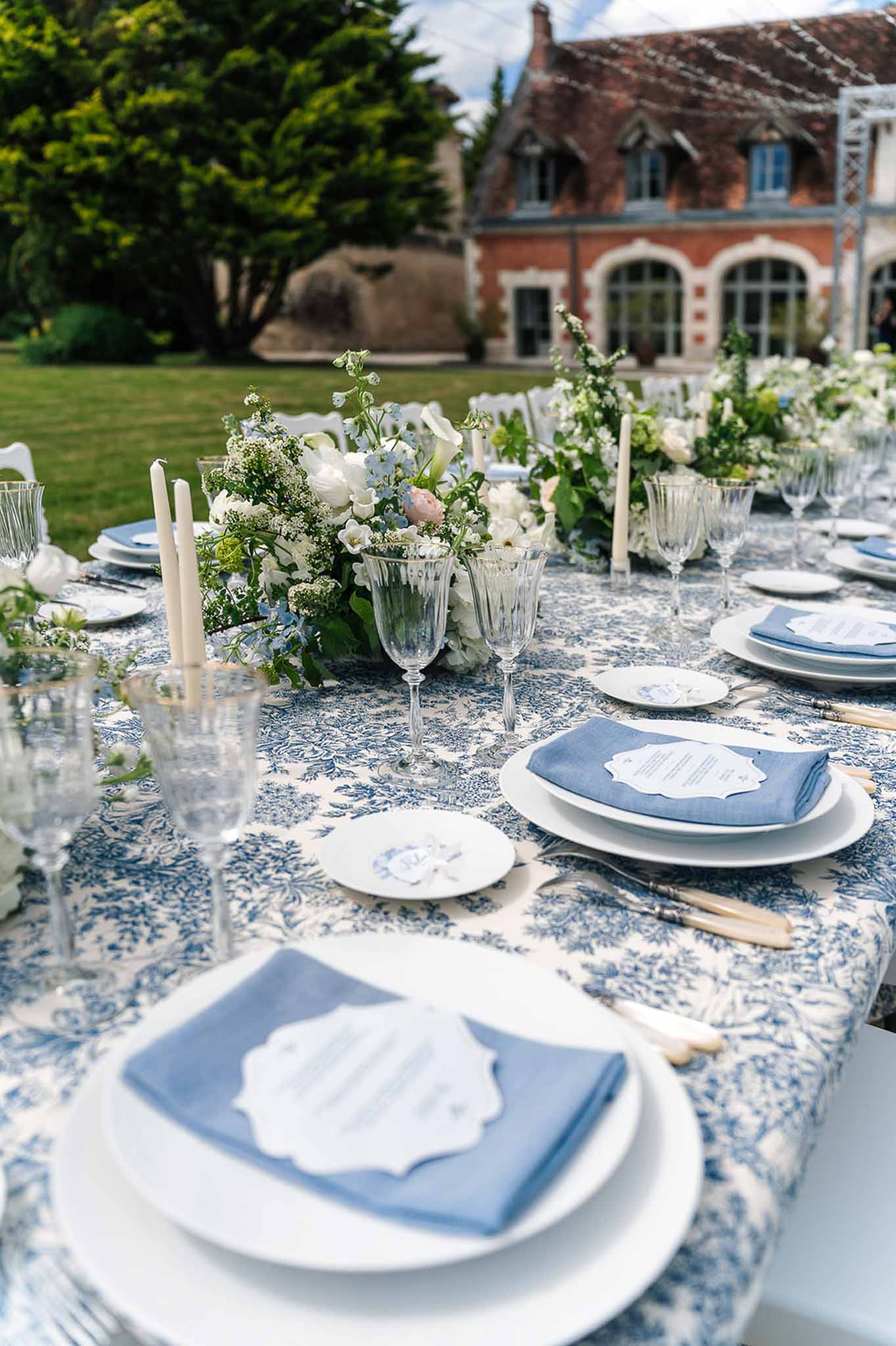 Long reception table with blue toile tablecloth, white ranunculus centerpieces, and taper candles at French manor