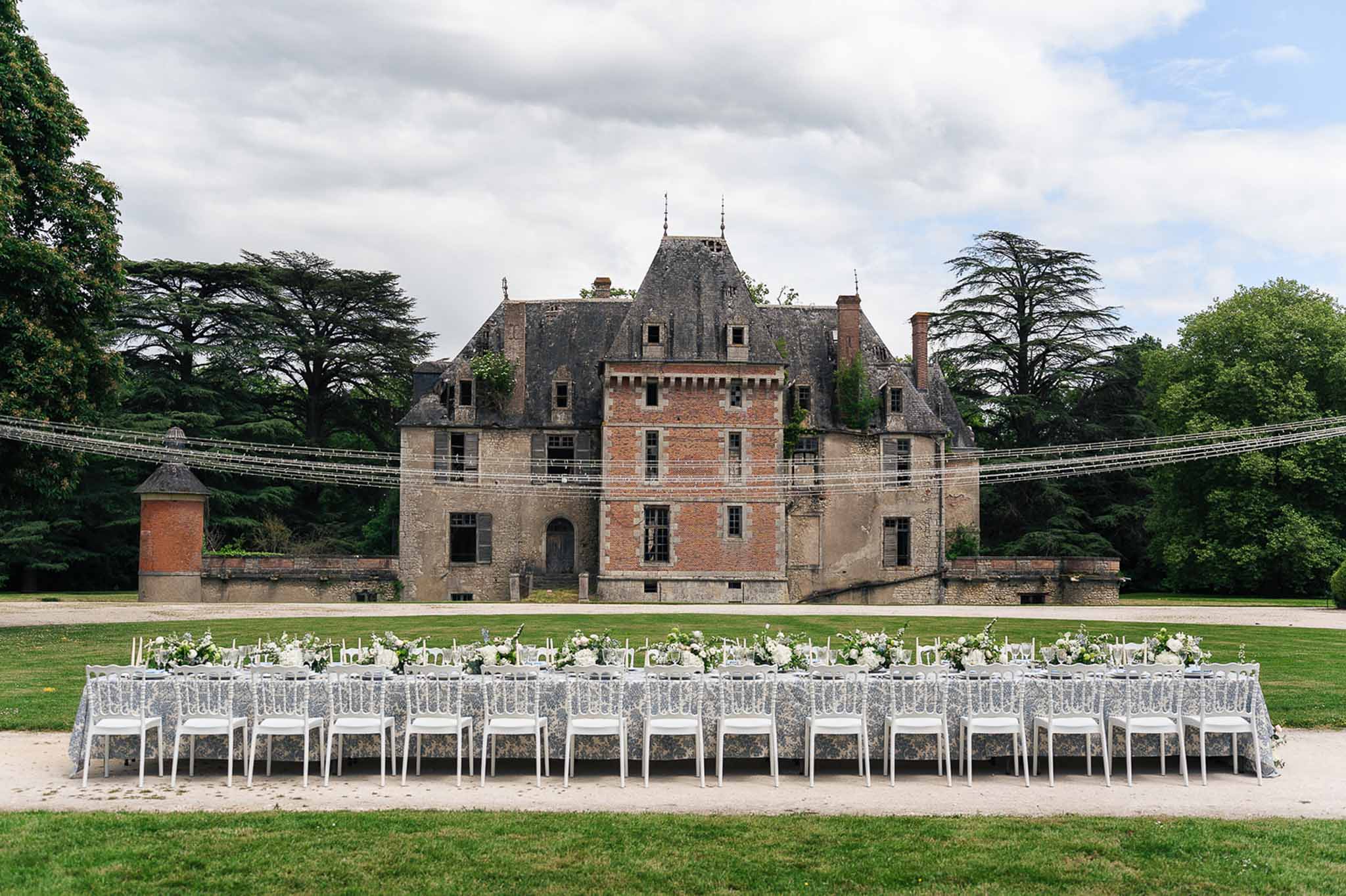 Long table with blue toile linen white roses ghost chairs and fairy light canopy before red-brick chateau