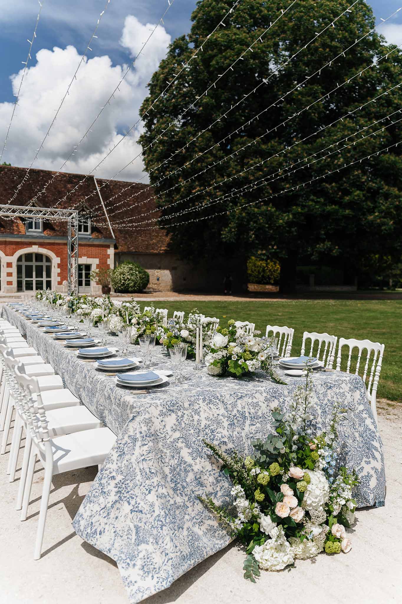 Long outdoor reception table with toile de Jouy cloth, white hydrangea floral runner, and fairy light canopy at French estate