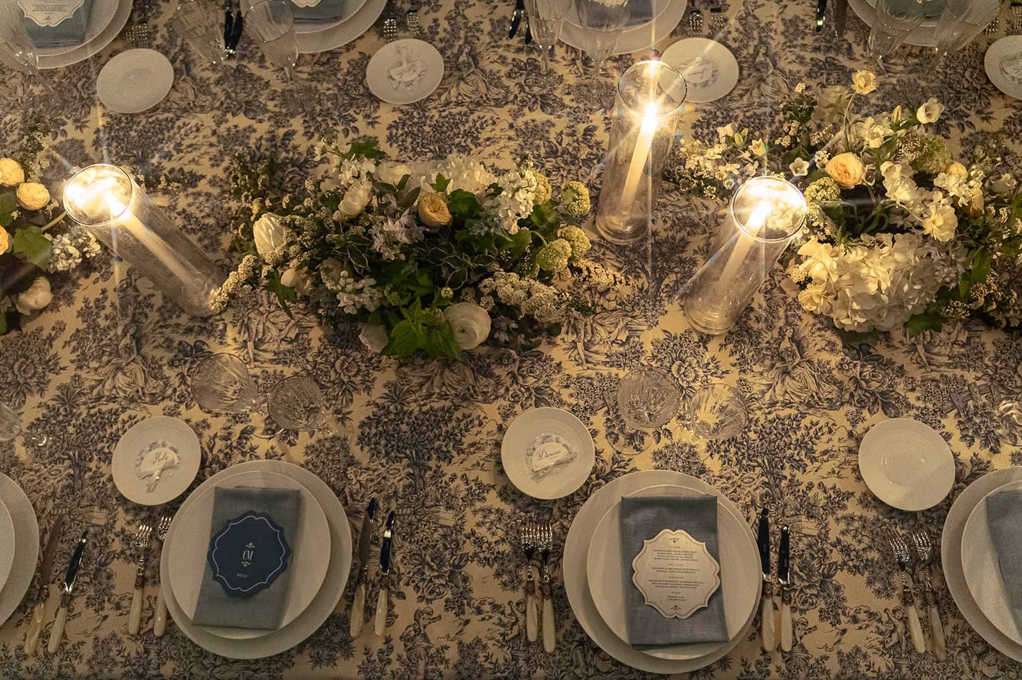Overhead view of reception table with navy toile de Jouy tablecloth, white ranunculus centerpieces, and hurricane candles