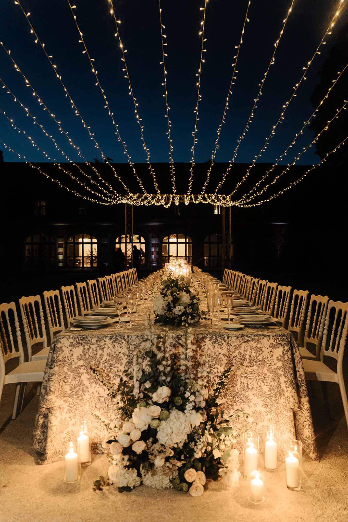 Evening courtyard reception table with floral jacquard linen gold chiavari chairs fairy light canopy and pillar candles