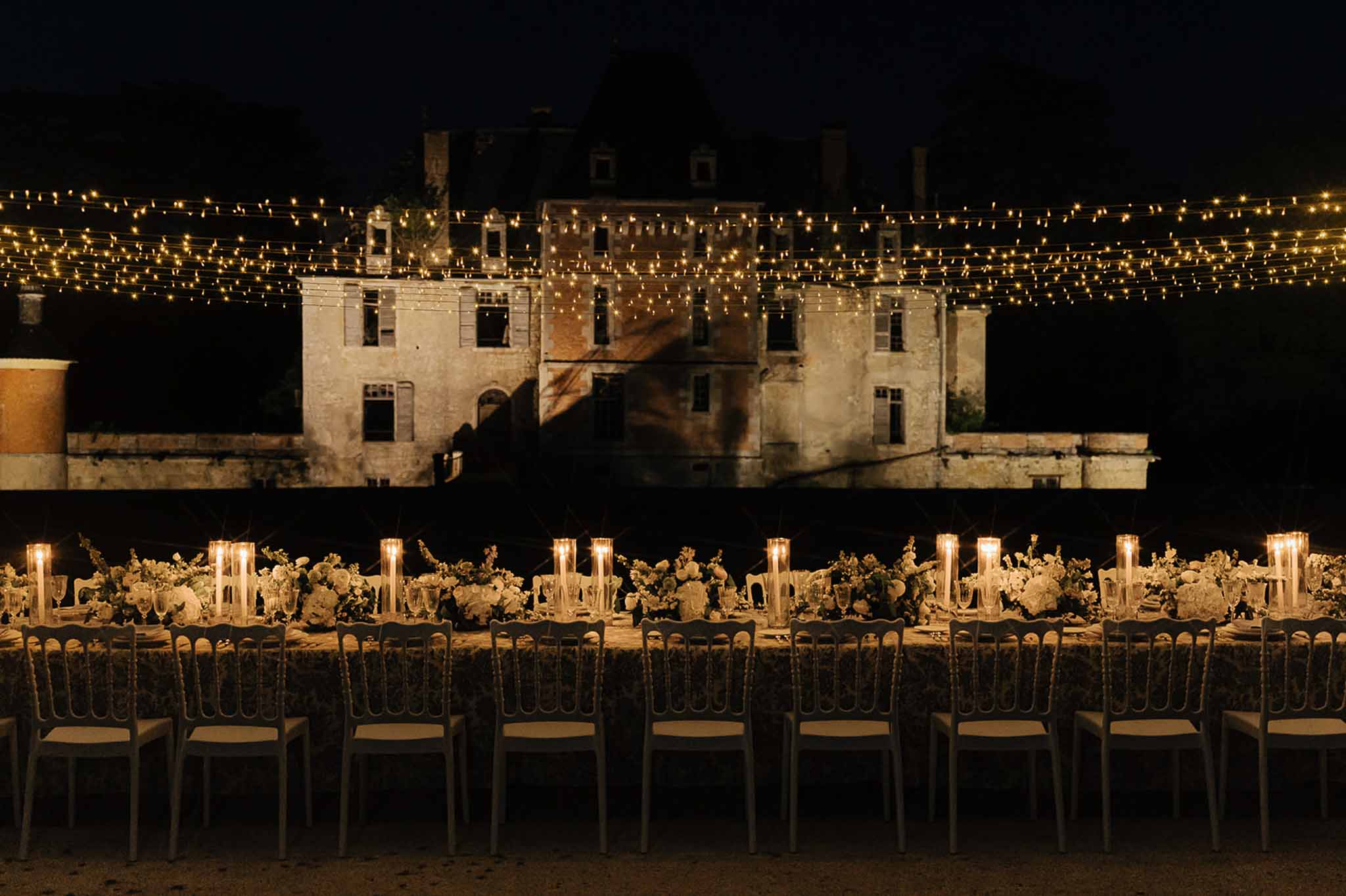 Long banquet table with white rose runner and pillar candles under fairy-light canopy before lit chateau