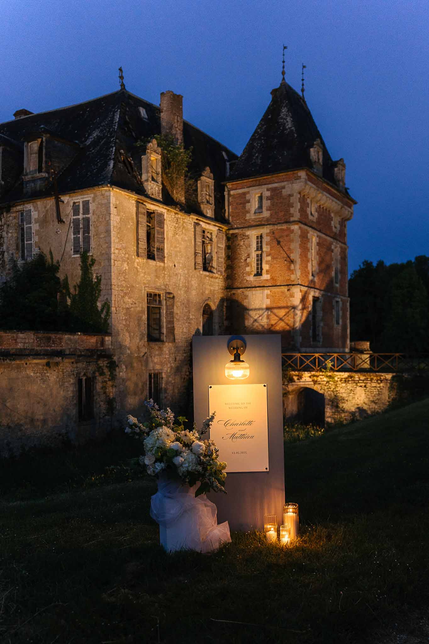 Gold-script welcome sign with white rose and blue accent arrangement and hurricane candles before lit chateau at dusk