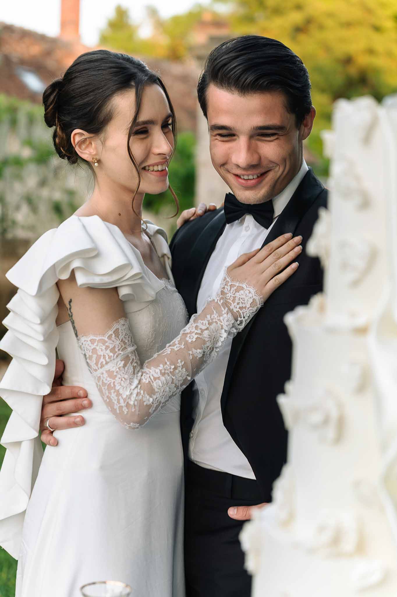Smiling bride in ruffled one-shoulder gown and groom in black tuxedo embracing beside white tiered cake
