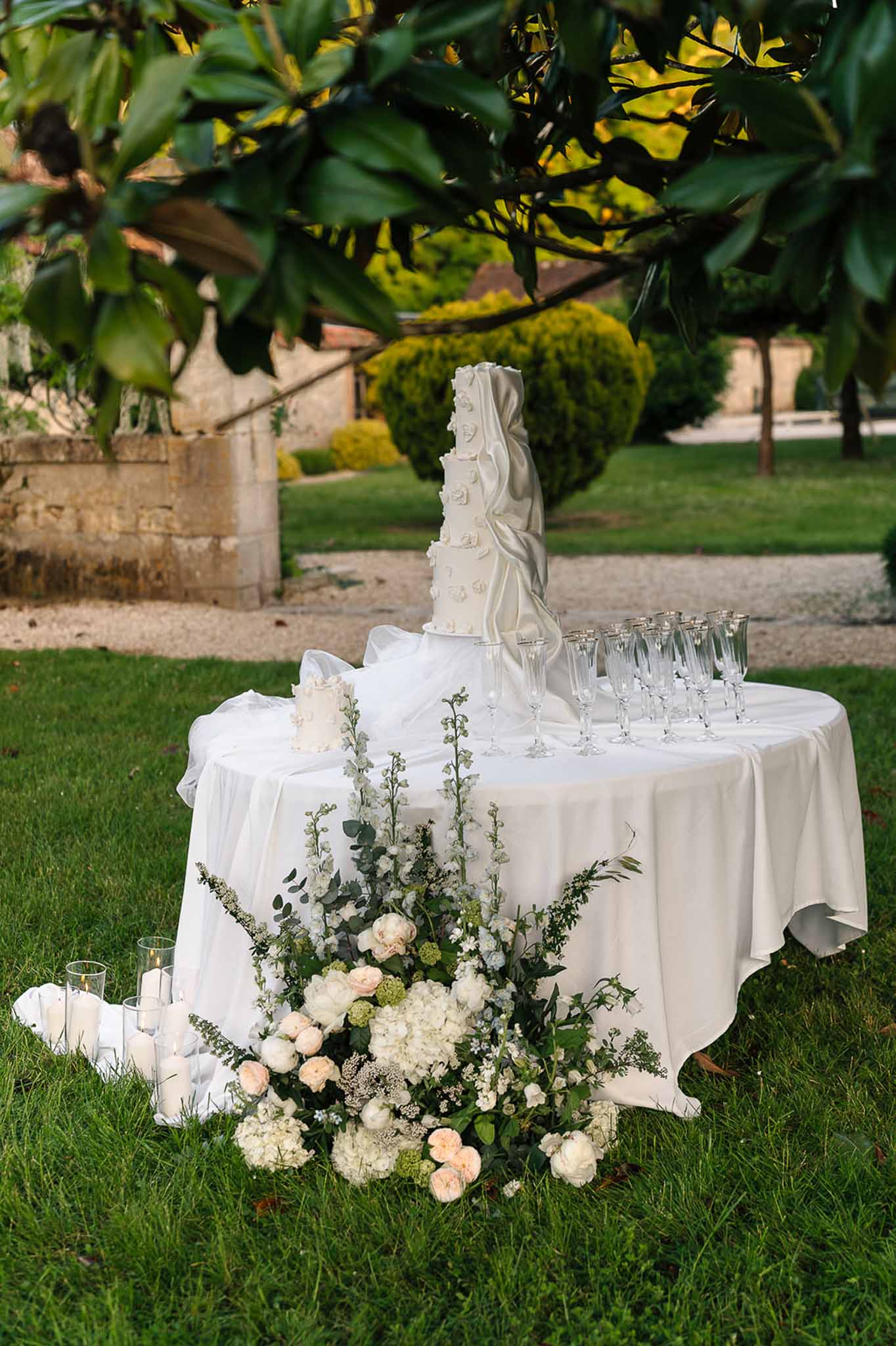 Multi-tier white wedding cake with champagne flutes and hydrangea arrangement on lawn
