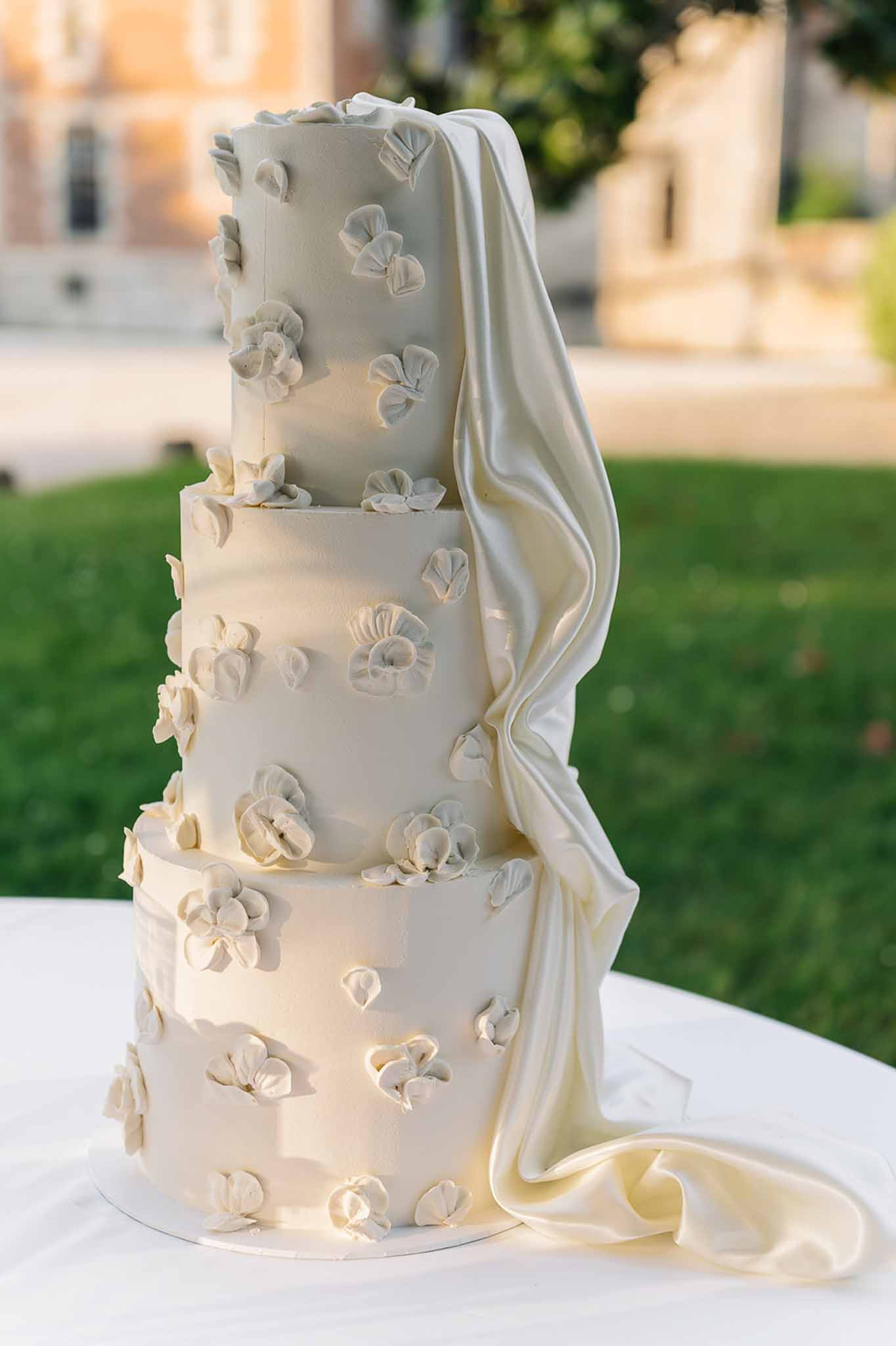 Four-tier ivory fondant cake with 3D sugar flower petals and draped satin ribbon on white linen with chateau behind