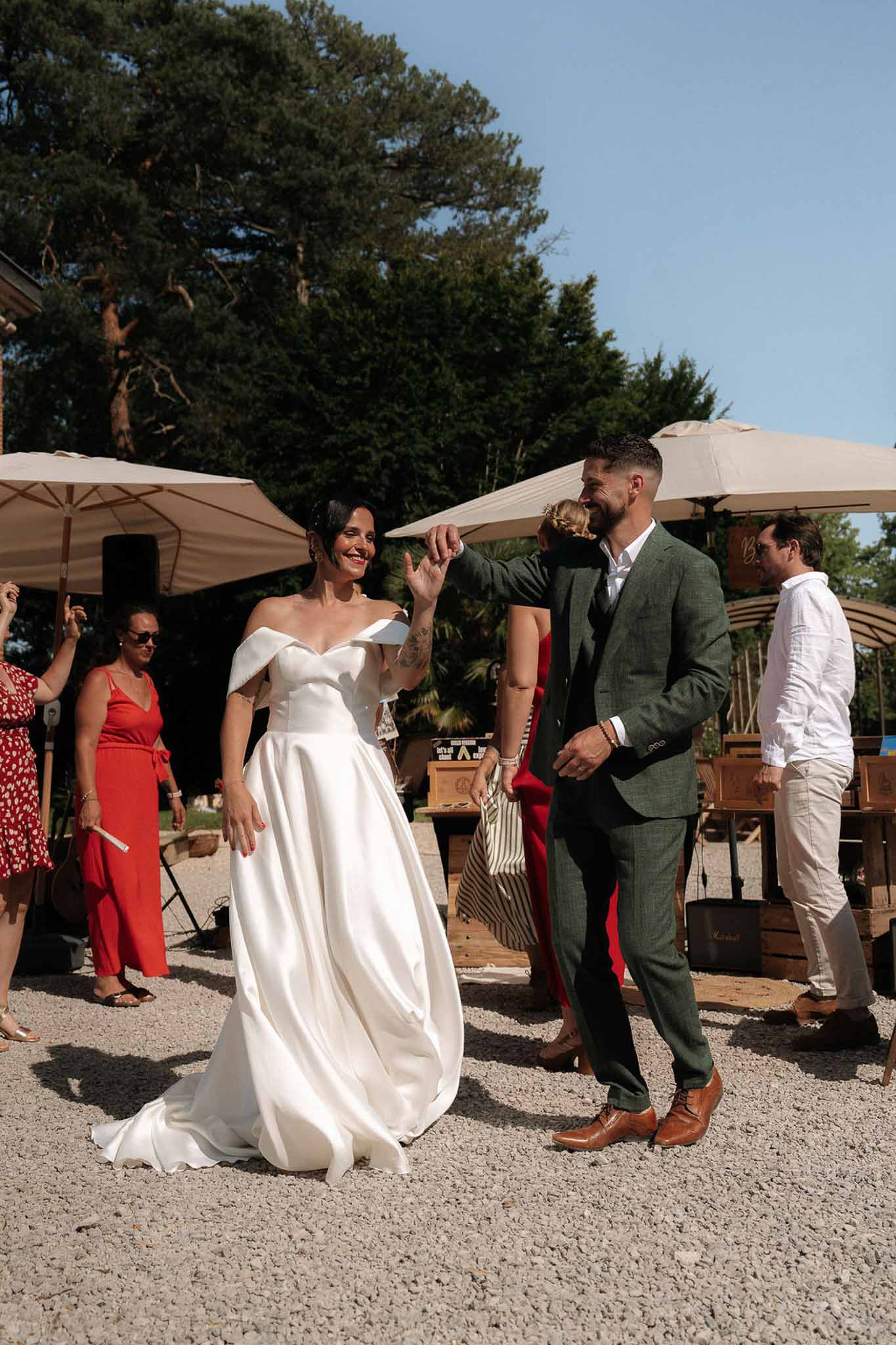 Bride in white off-the-shoulder ballgown spinning with groom in green tweed suit on gravel courtyard