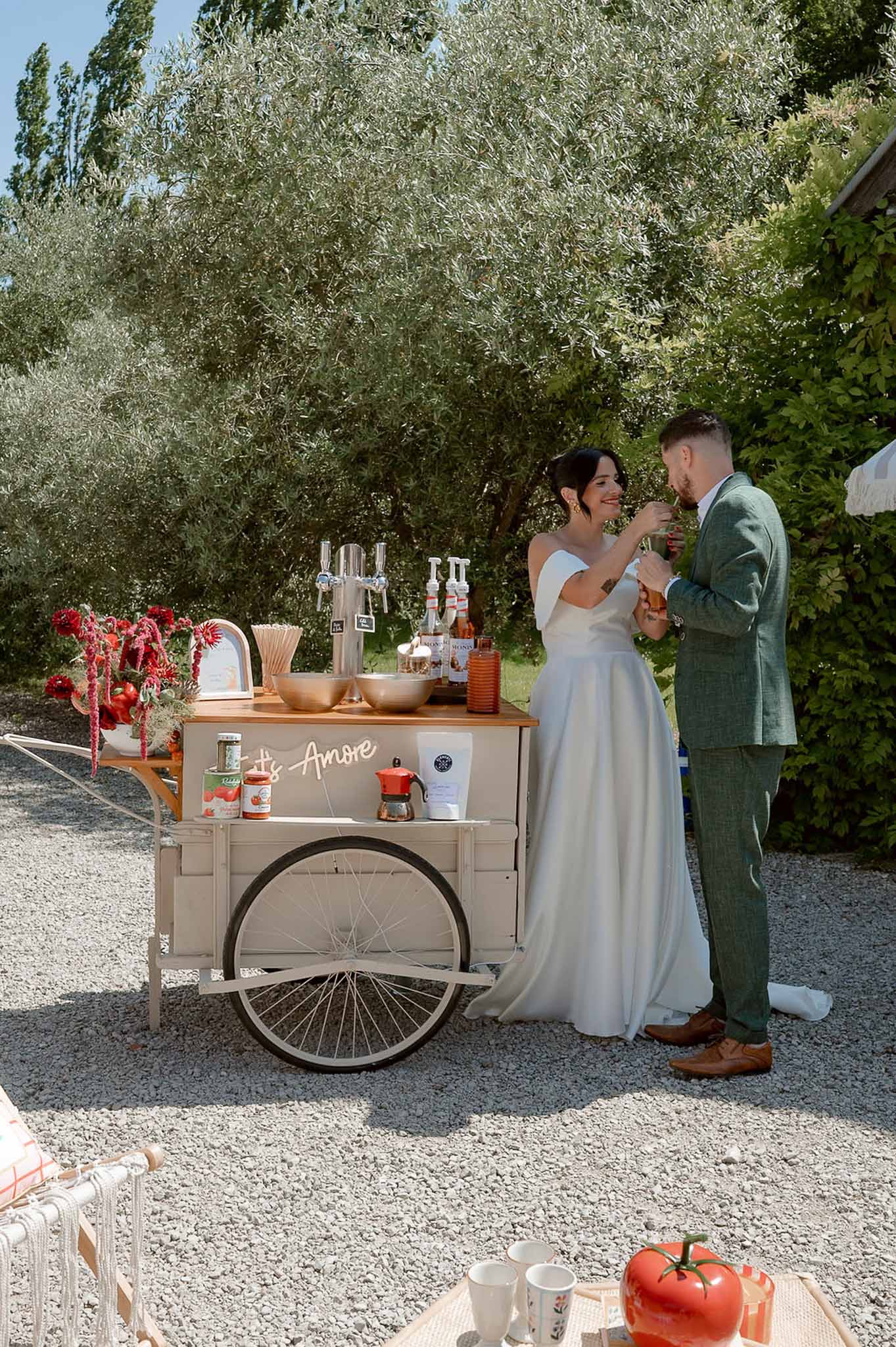 Bride in white gown and groom in sage suit at vintage cocktail cart with neon sign under olive trees