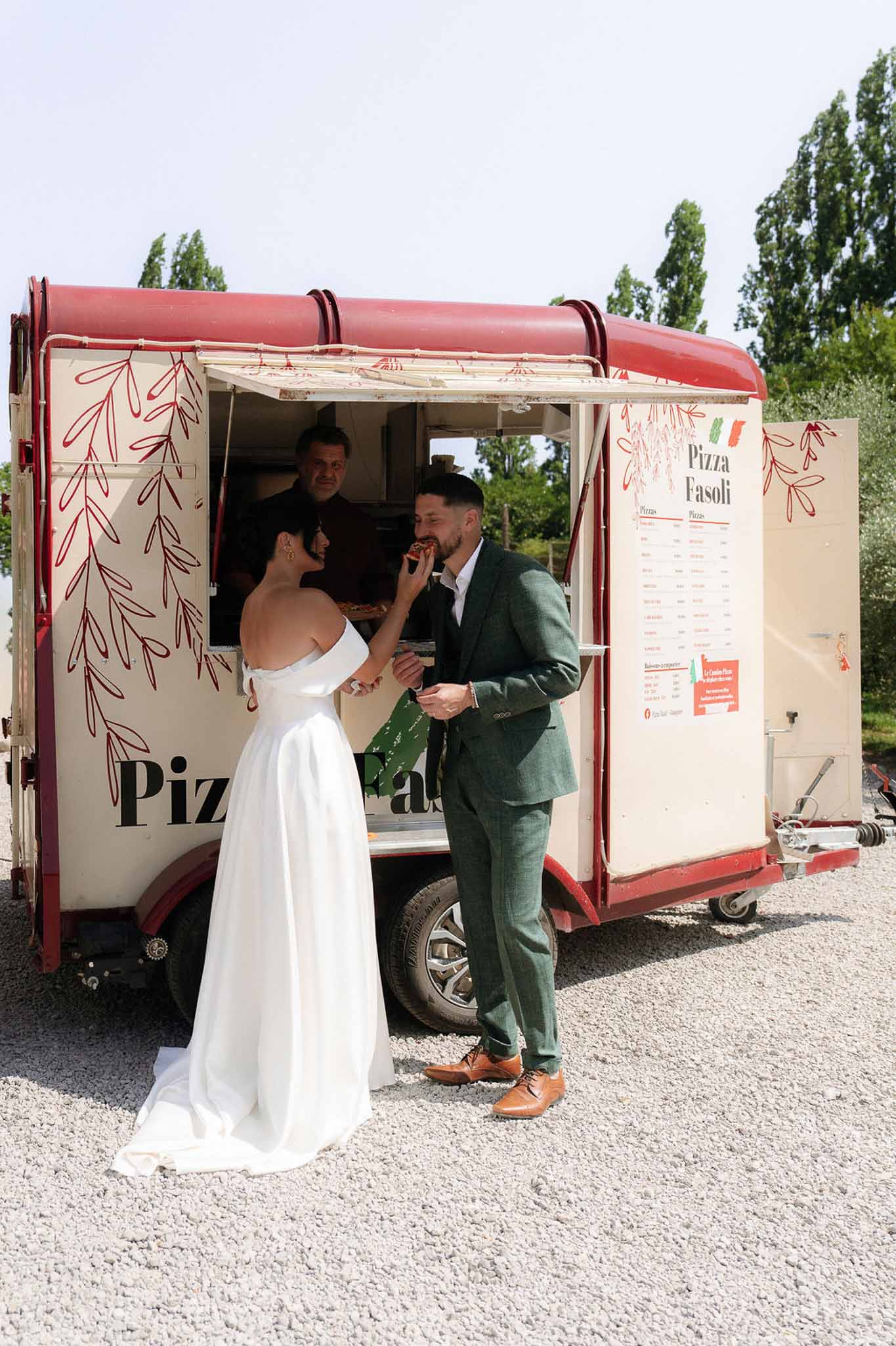 Bride feeding groom pizza at cream and red food truck, bride in white satin gown, groom in green tweed suit