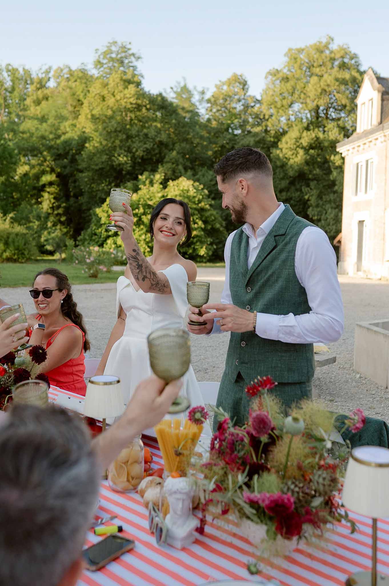 Bride and groom toasting with green goblets at eclectic table with crimson dahlias and striped cloth
