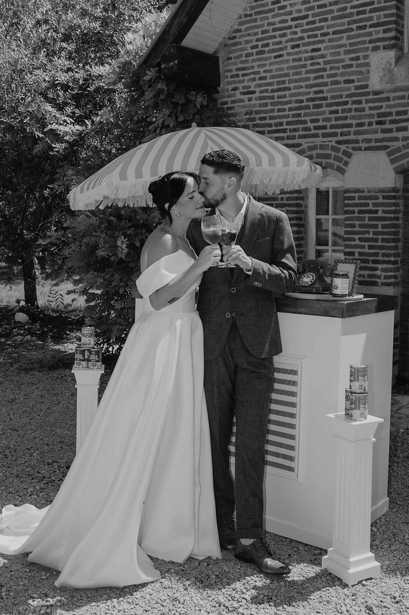 Bride and groom clinking wine glasses and kissing at outdoor cocktail bar in black and white
