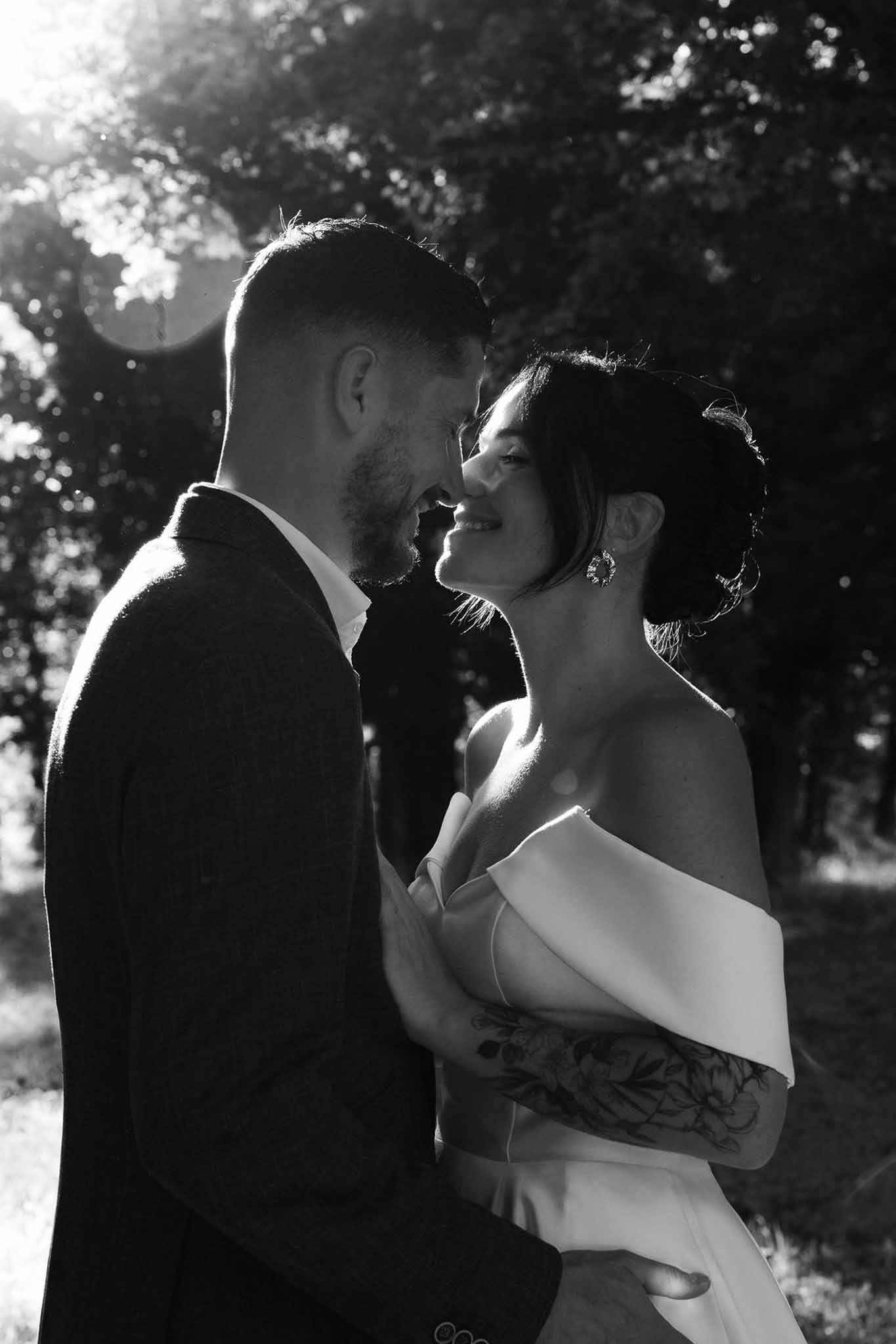 Black and white backlit portrait of bride and groom with foreheads touching, bride smiling upward