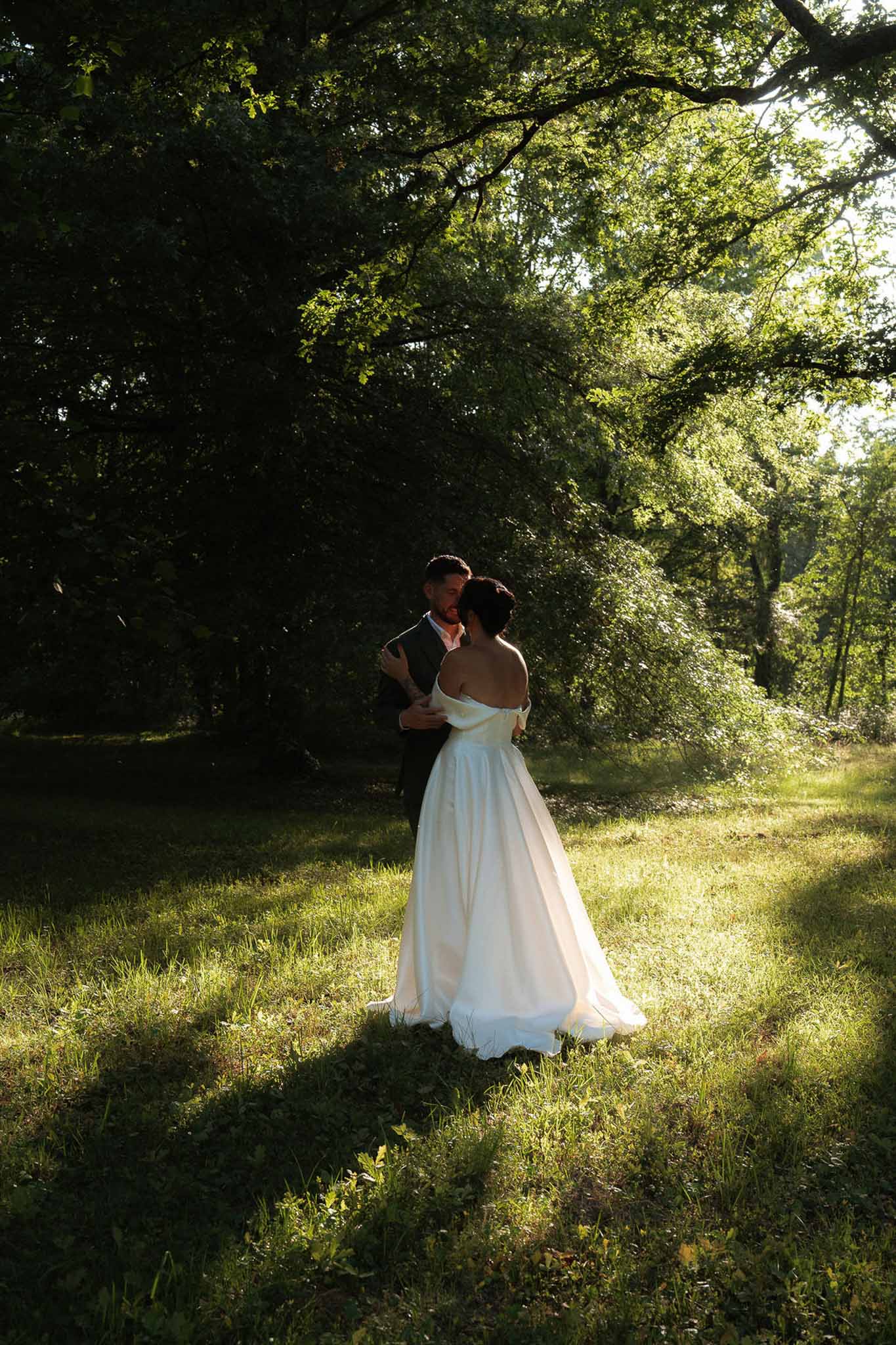 Couple embracing in wooded clearing during golden hour, bride in off-shoulder ballgown, warm backlight through trees