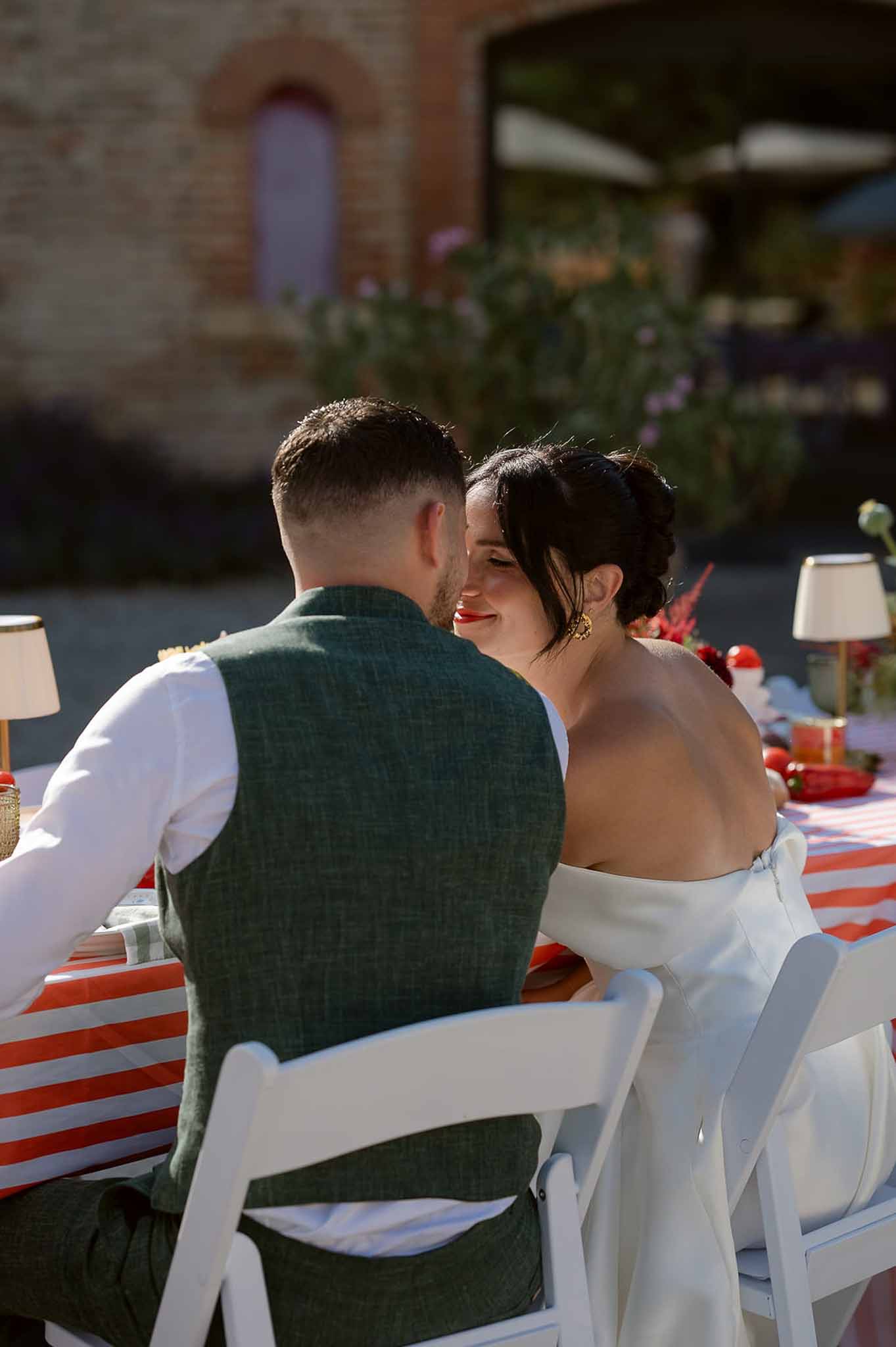 Bride and groom seated together at outdoor reception table with red and white striped runner and gold lamps