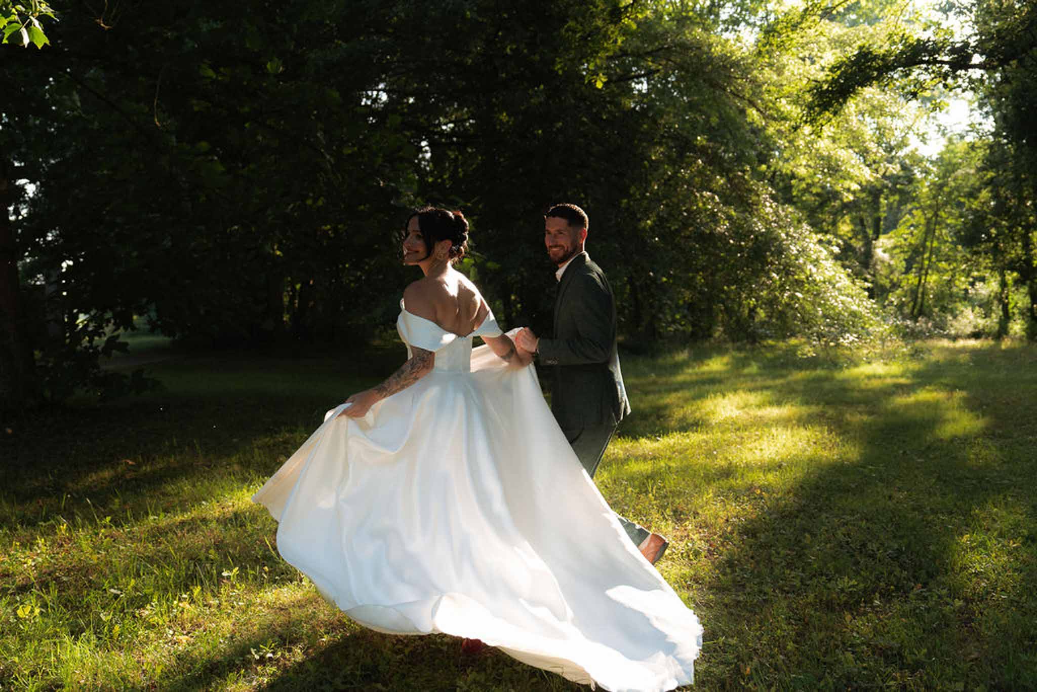 Couple walking through dappled woodland at golden hour, bride in off-shoulder ballgown, groom in green suit
