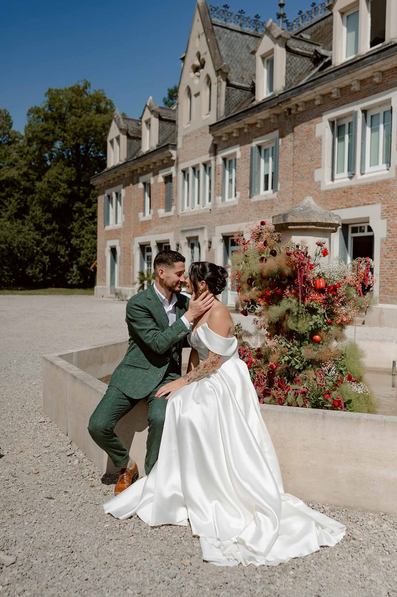 Bride and groom at stone fountain with autumnal floral arrangement in front of brick chateau