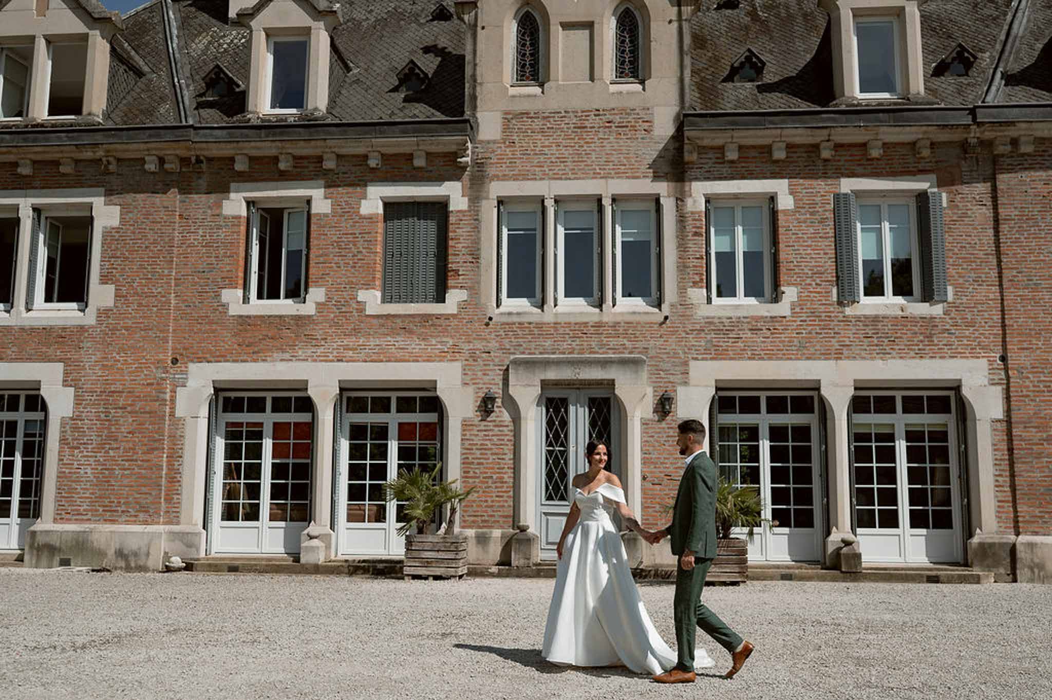Bride in white ballgown and groom in green suit walking hand in hand on chateau gravel courtyard