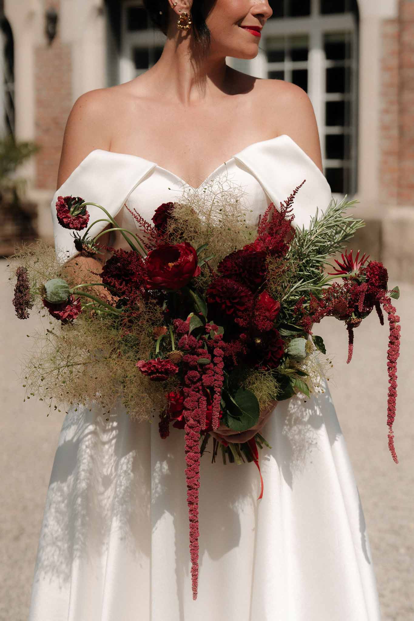 Bride in white off-shoulder gown holding oversized crimson and burgundy bouquet with peonies and trailing amaranthus