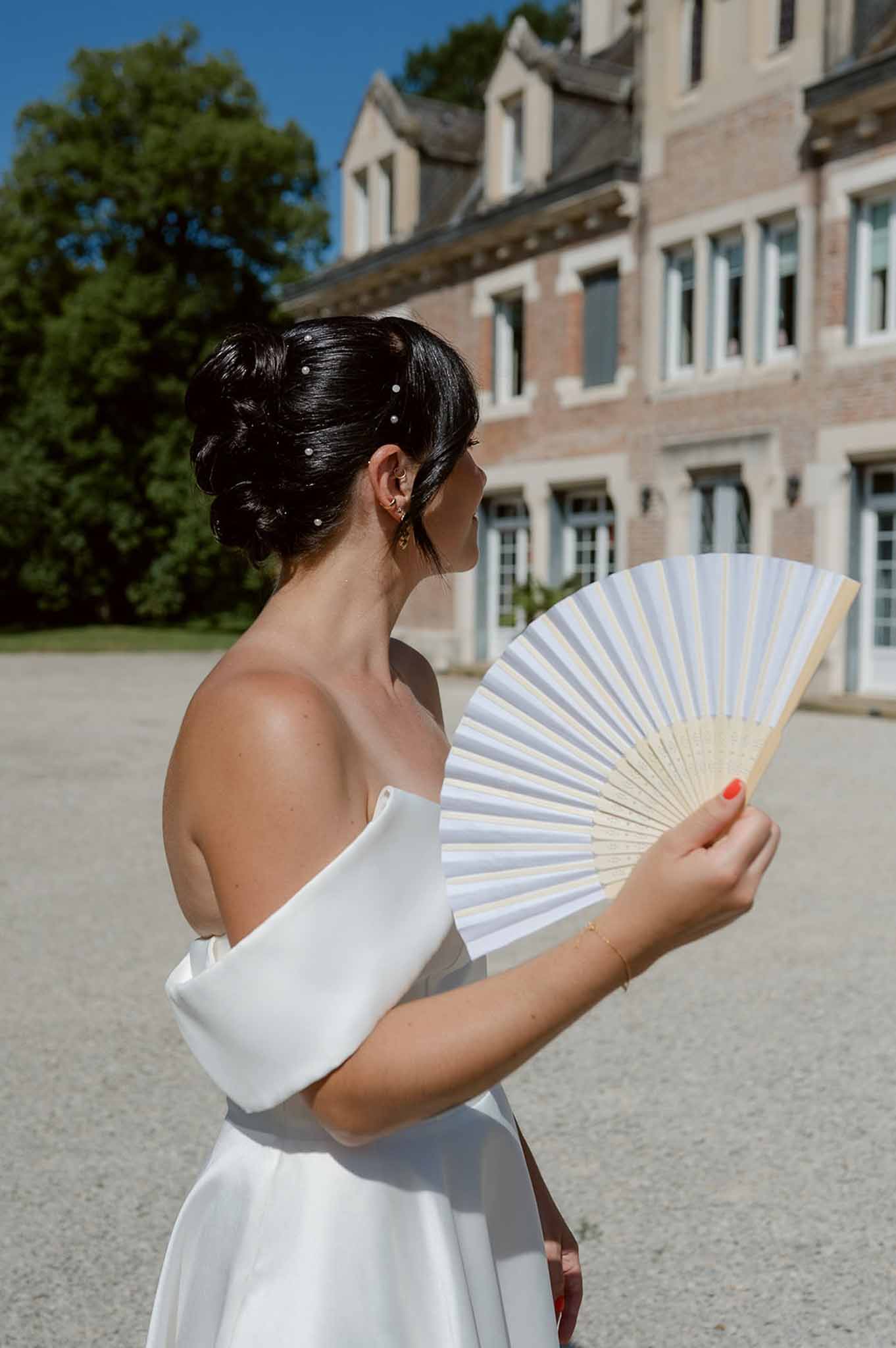 Bride in profile wearing off-shoulder white gown holding white hand fan in chateau courtyard with pearl hair pins and gold...