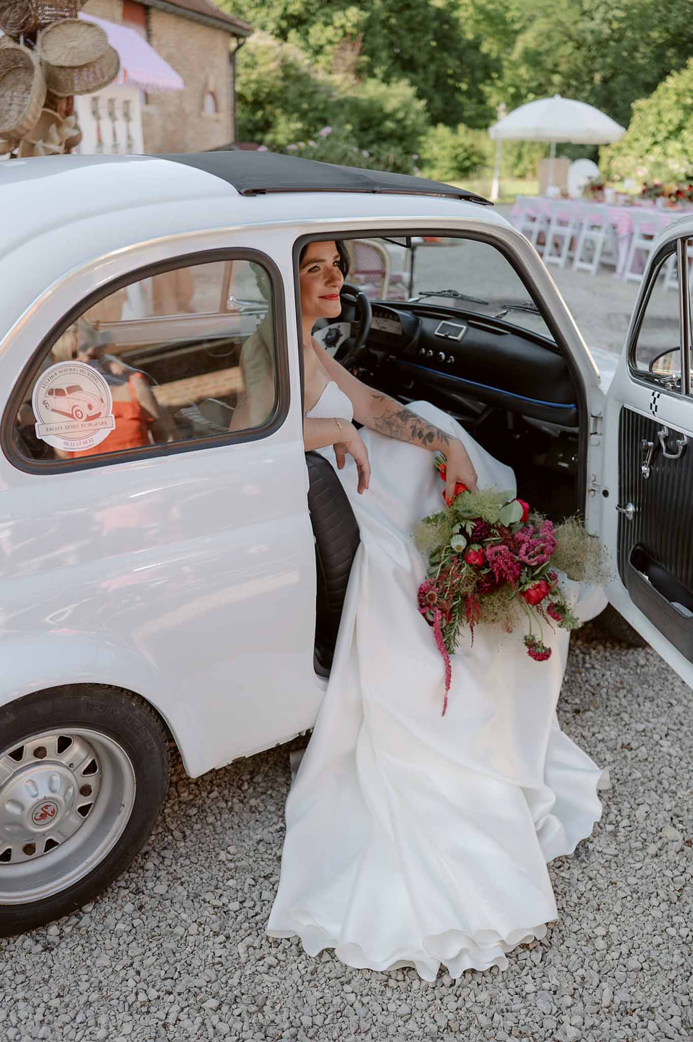 Bride in white ballgown holding magenta and red anemone bouquet seated in vintage white Citroen 2CV
