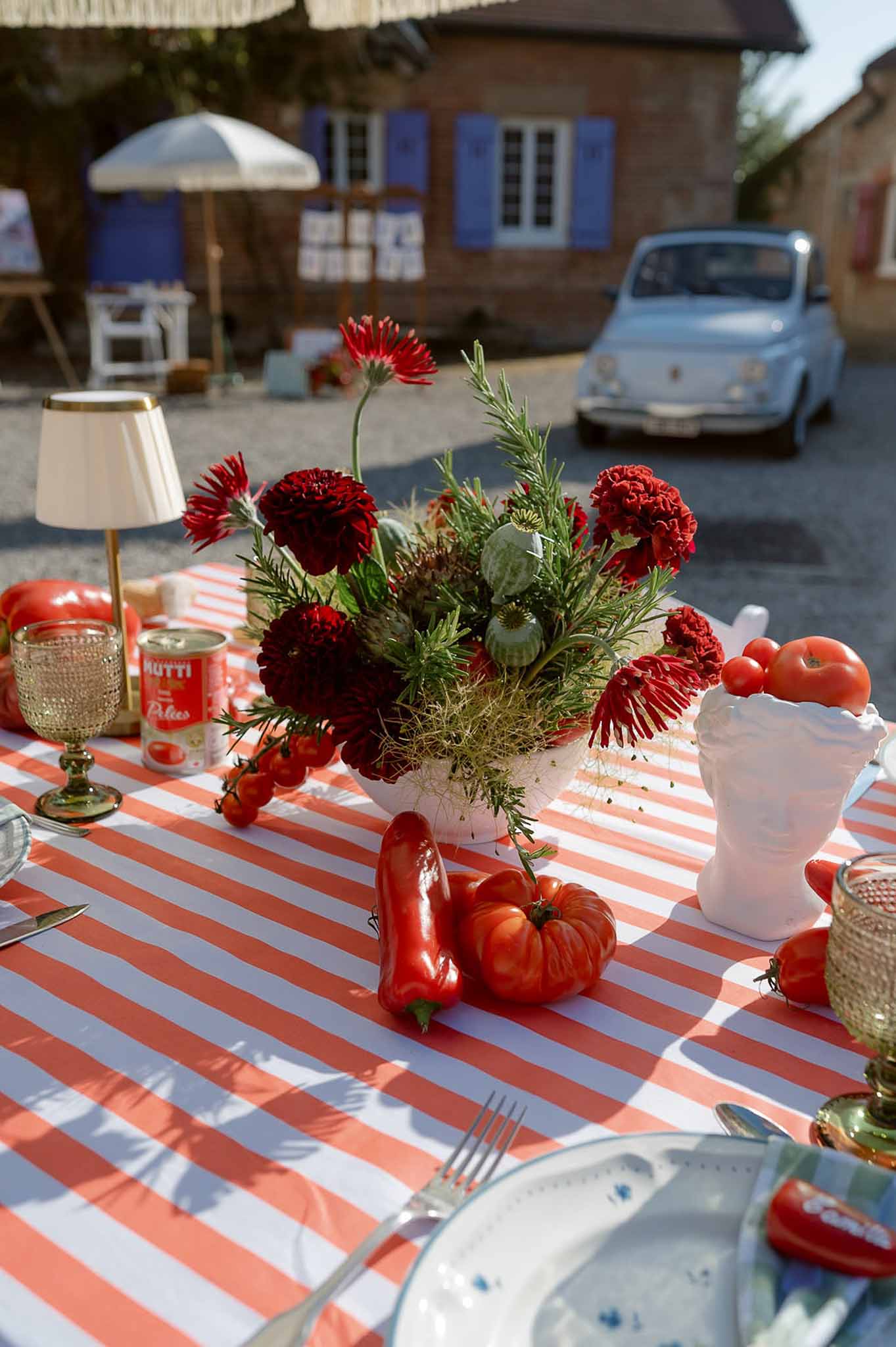 Red dahlia centerpiece with tomatoes and green glassware on striped tablecloth with blue vintage Fiat in courtyard
