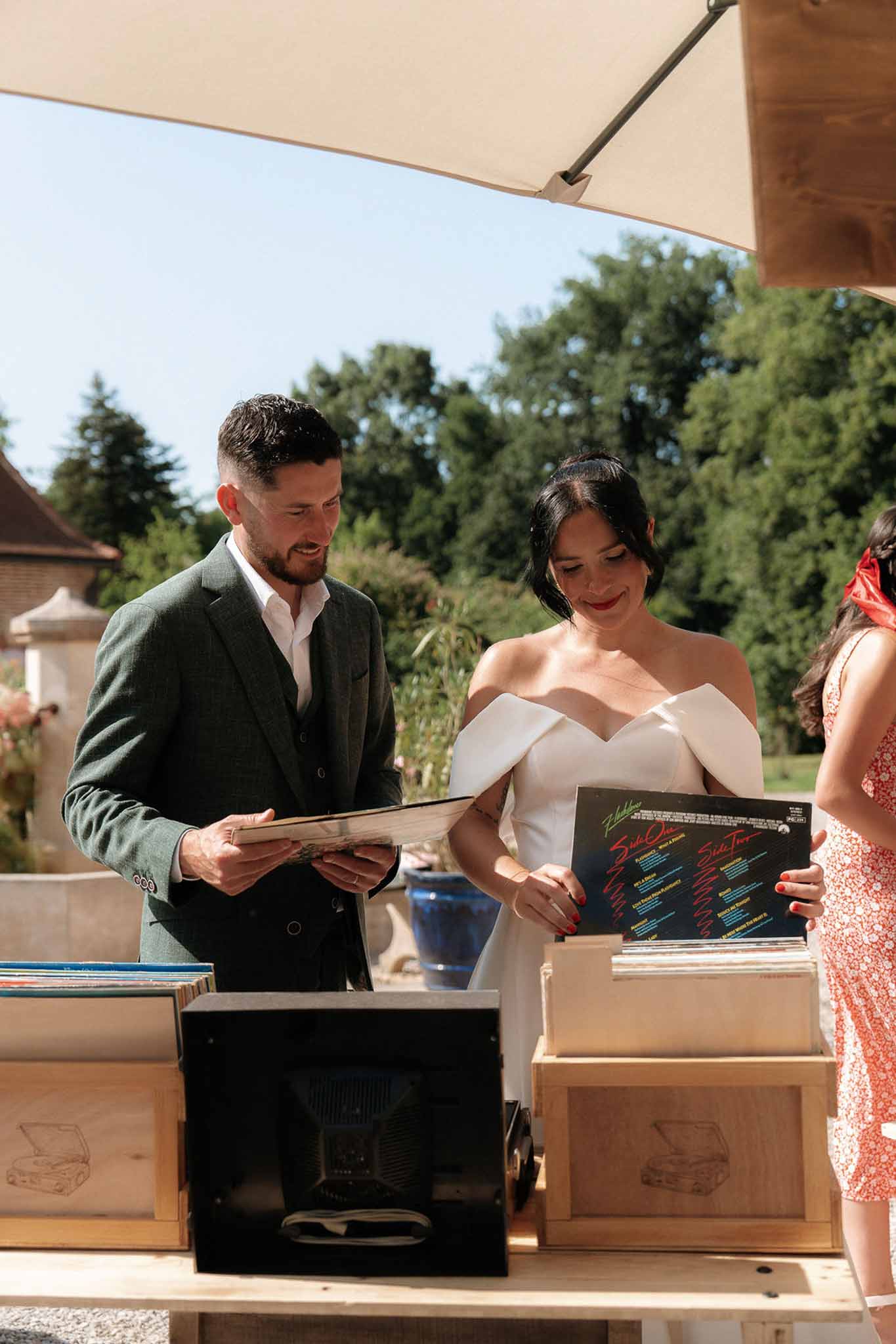 Couple browsing vinyl records at outdoor station under cream umbrella, groom in green tweed, bride in white gown
