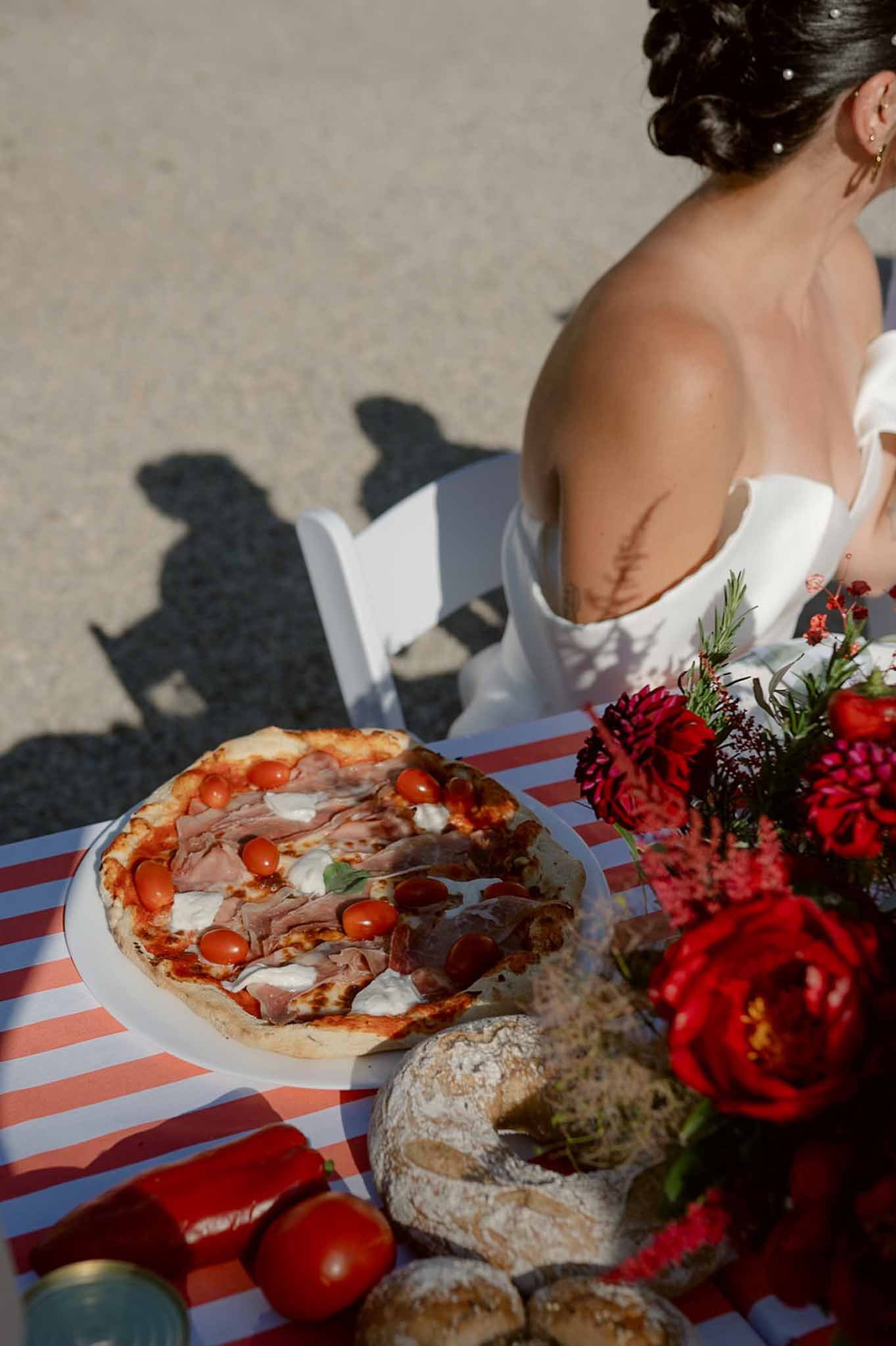 Pizza on red-striped tablecloth with burgundy ranunculus bouquet and bride in off-shoulder gown behind