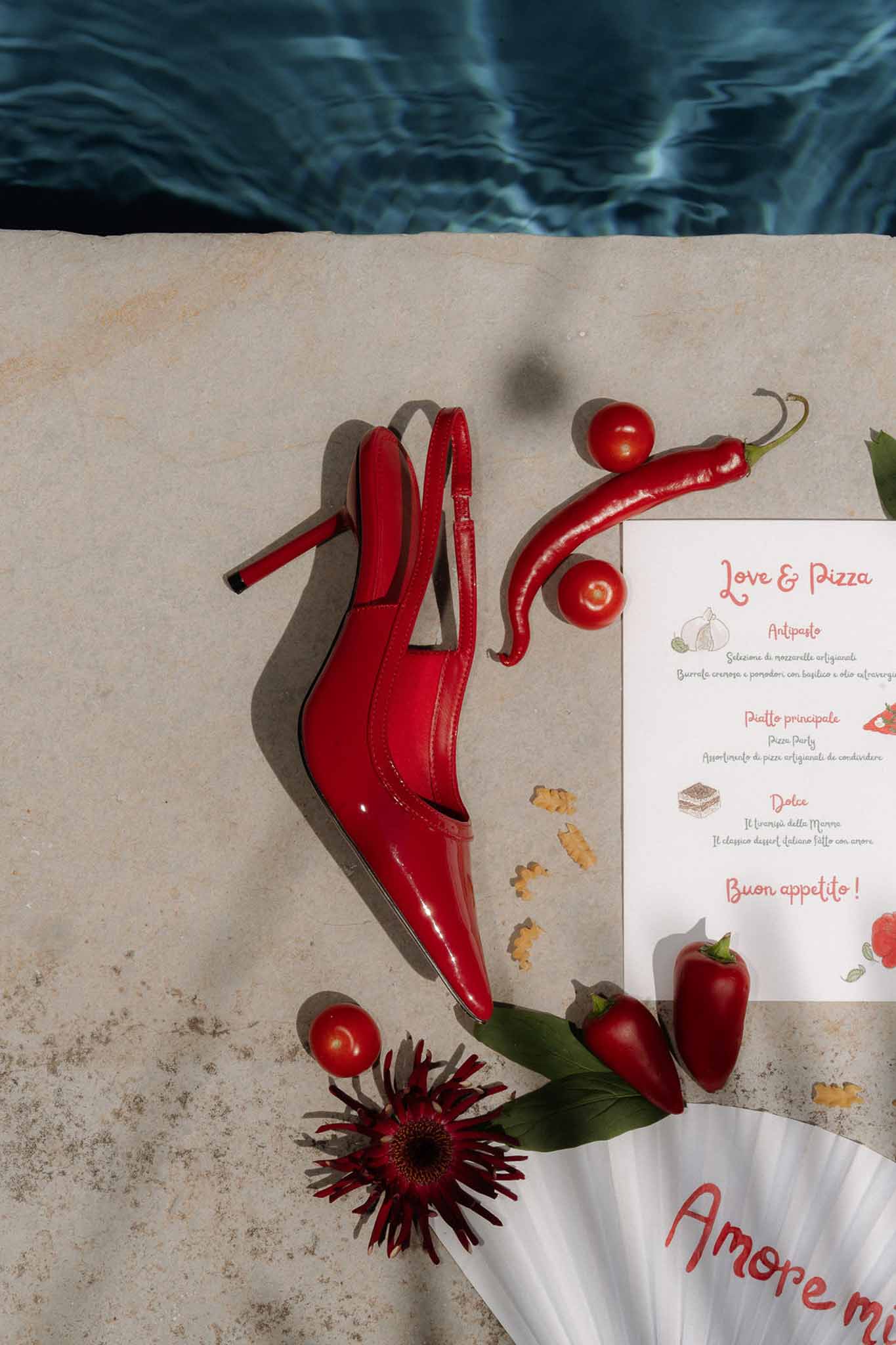 Red patent slingback heel with Love and Pizza menu card, tomatoes, and chili peppers by pool