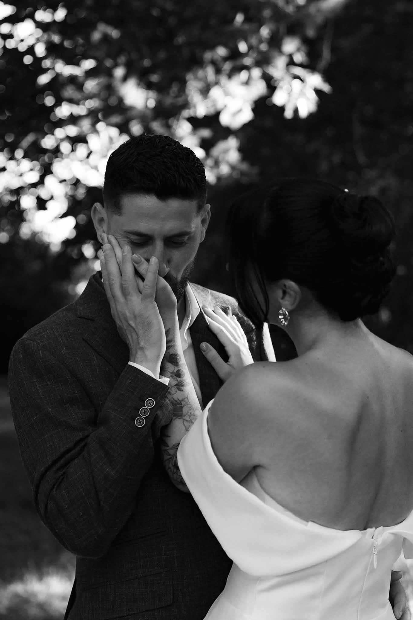 Groom covering mouth emotionally as bride rests hand on his chest during outdoor first look in B&W