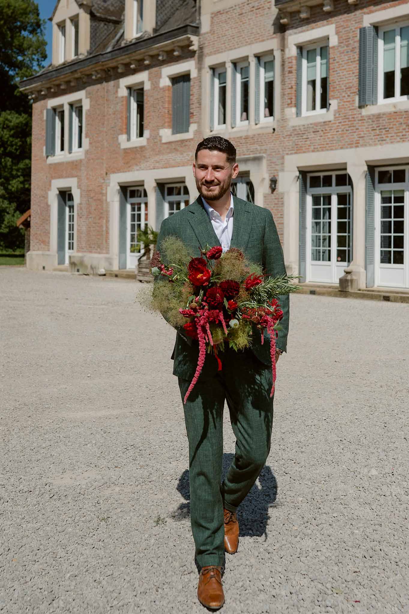 Groom in forest green suit carrying crimson peony and amaranthus bouquet across chateau gravel courtyard