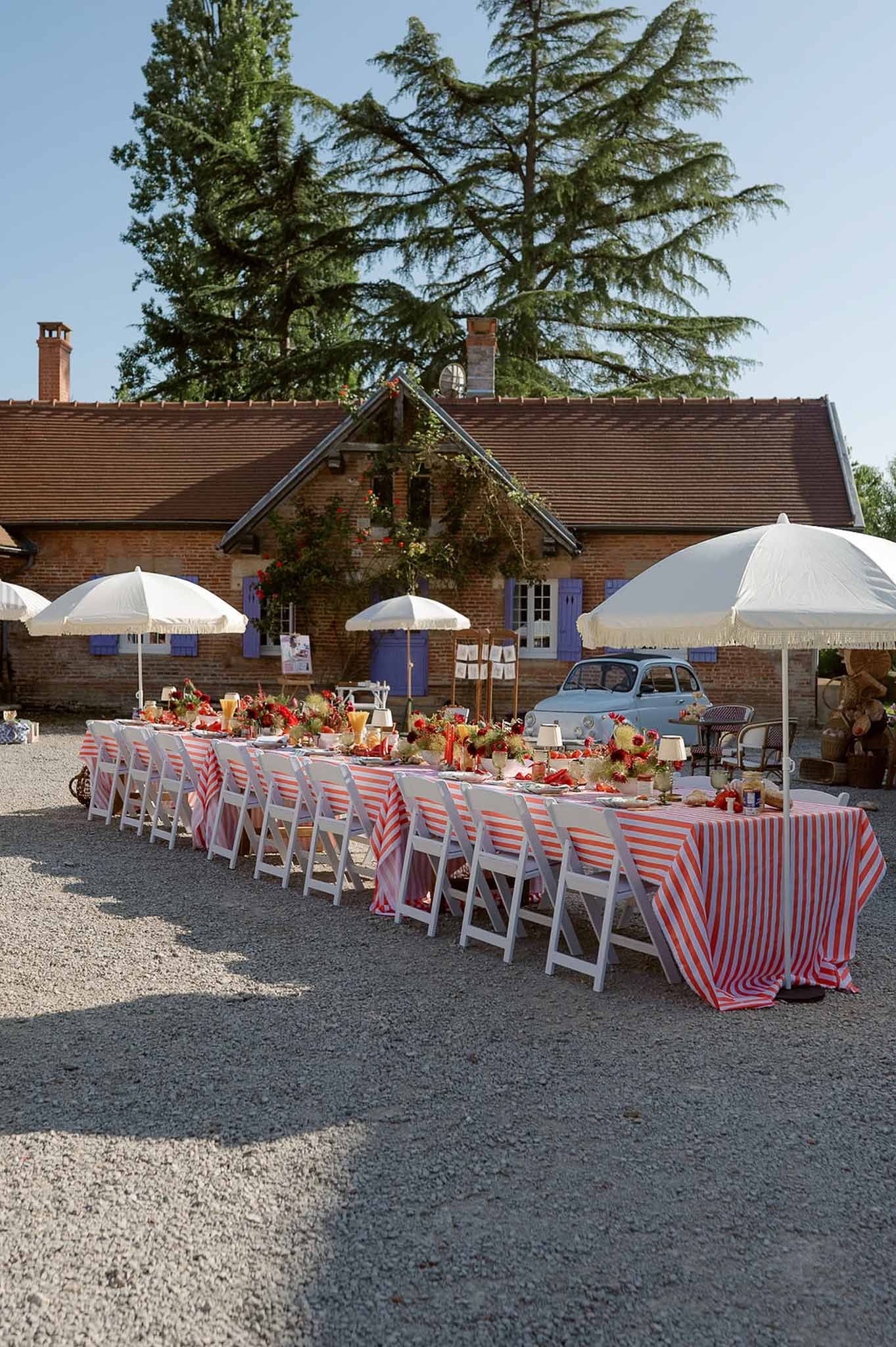 Long reception table with orange striped tablecloth and red-orange centerpieces on gravel courtyard with white umbrellas