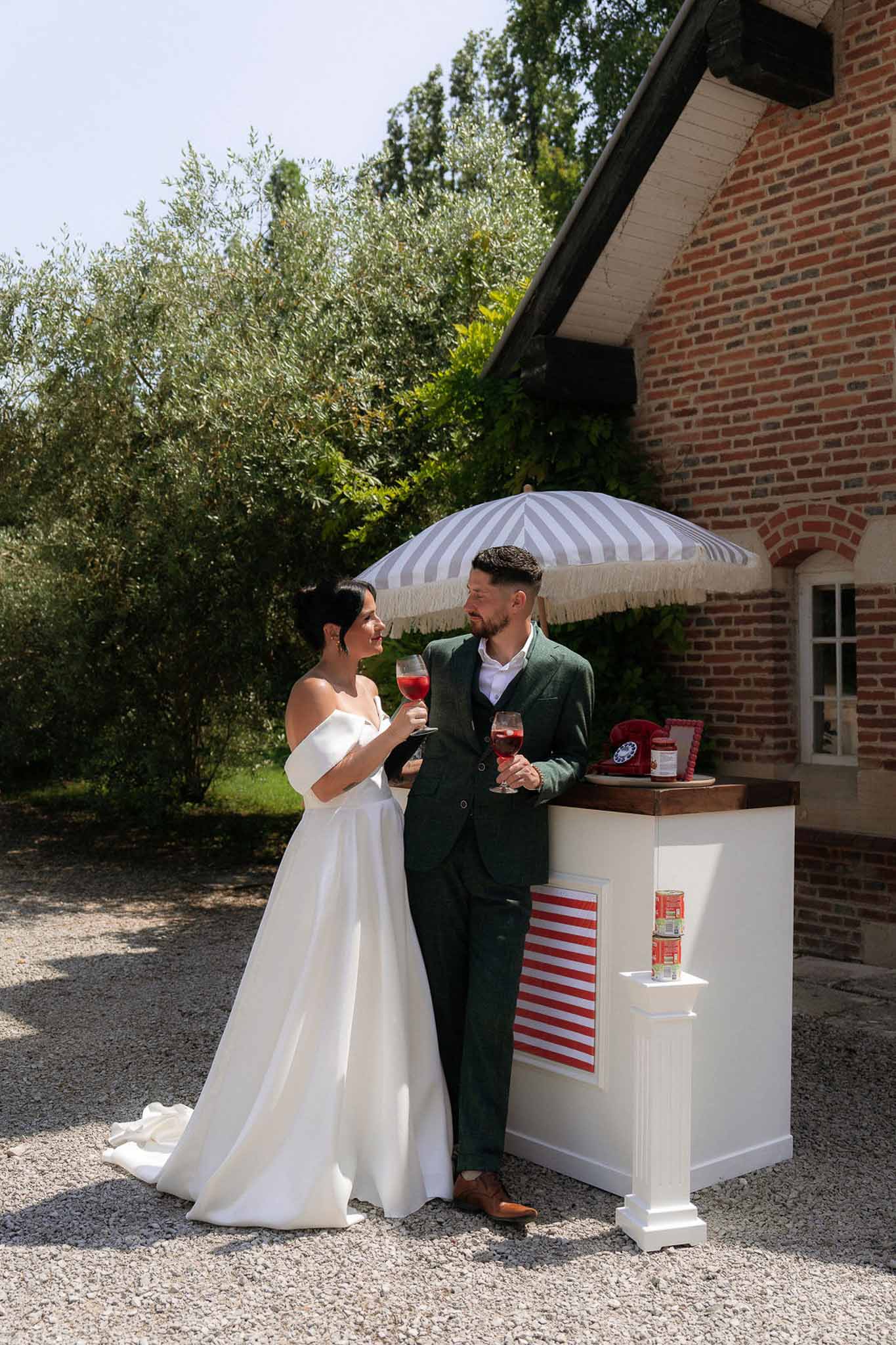 Bride and groom holding drinks at retro-styled outdoor cocktail bar with striped parasol