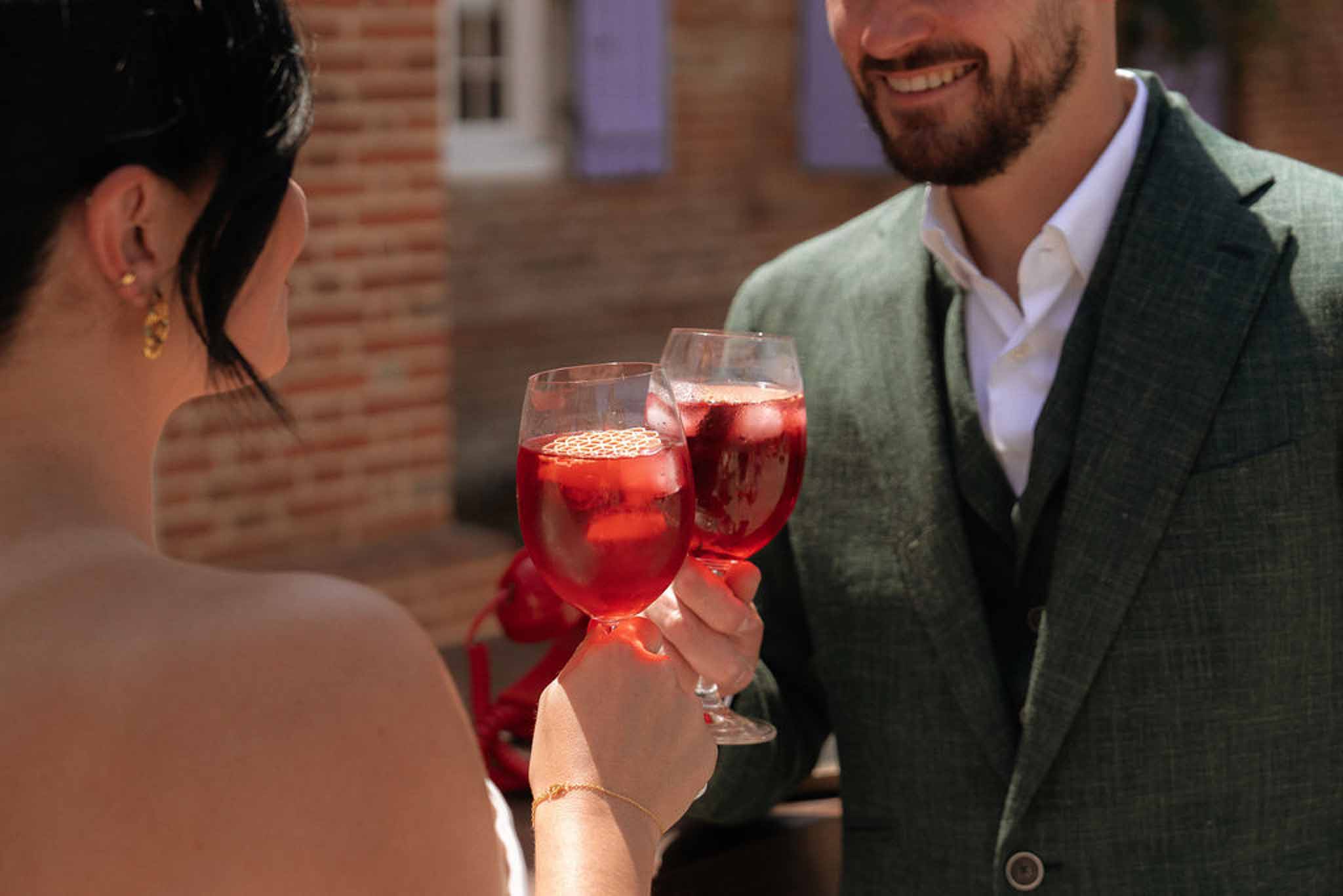 Couple clinking red cocktail glasses during outdoor cocktail hour at a brick venue with lavender shutters