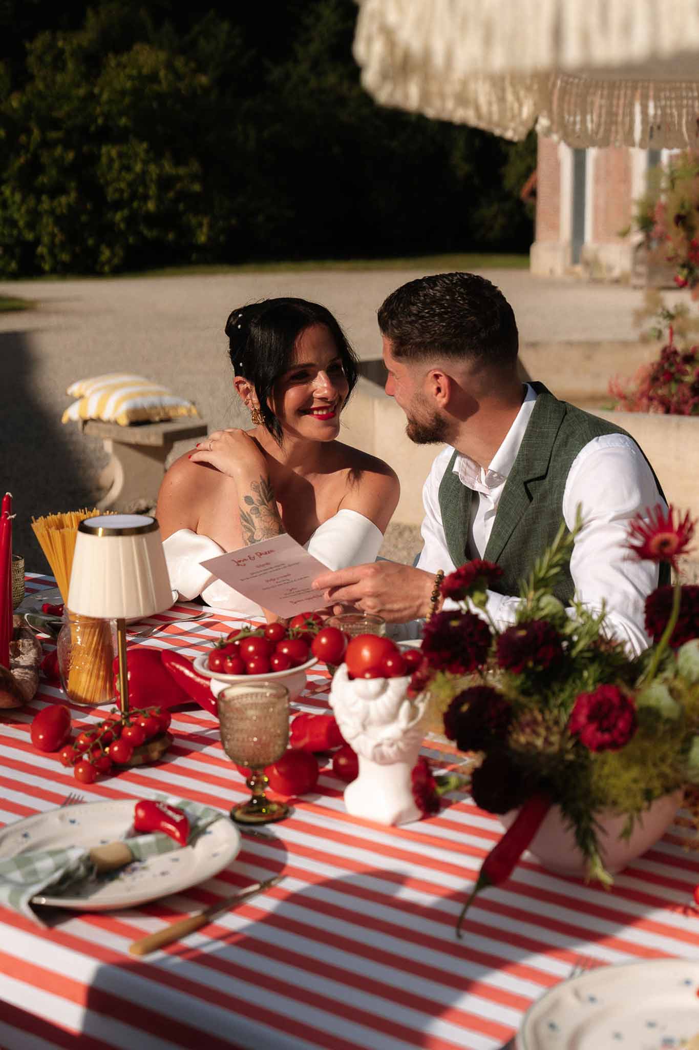 Couple at Italian-themed sweetheart table with red stripe linen, tomatoes, and burgundy dahlia decor