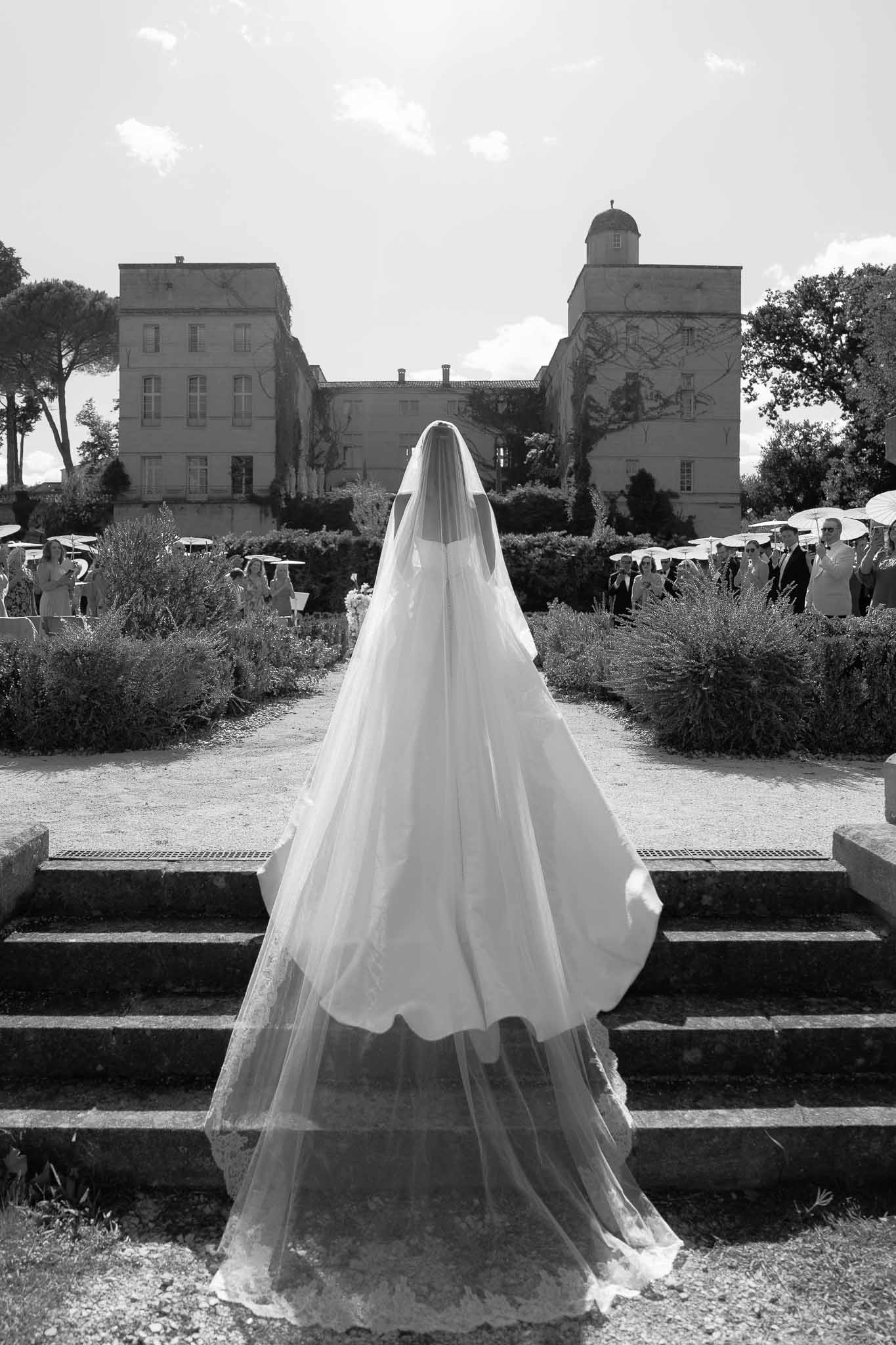 Black and white bride from behind with cathedral veil on stone steps as guests hold parasols before domed chateau