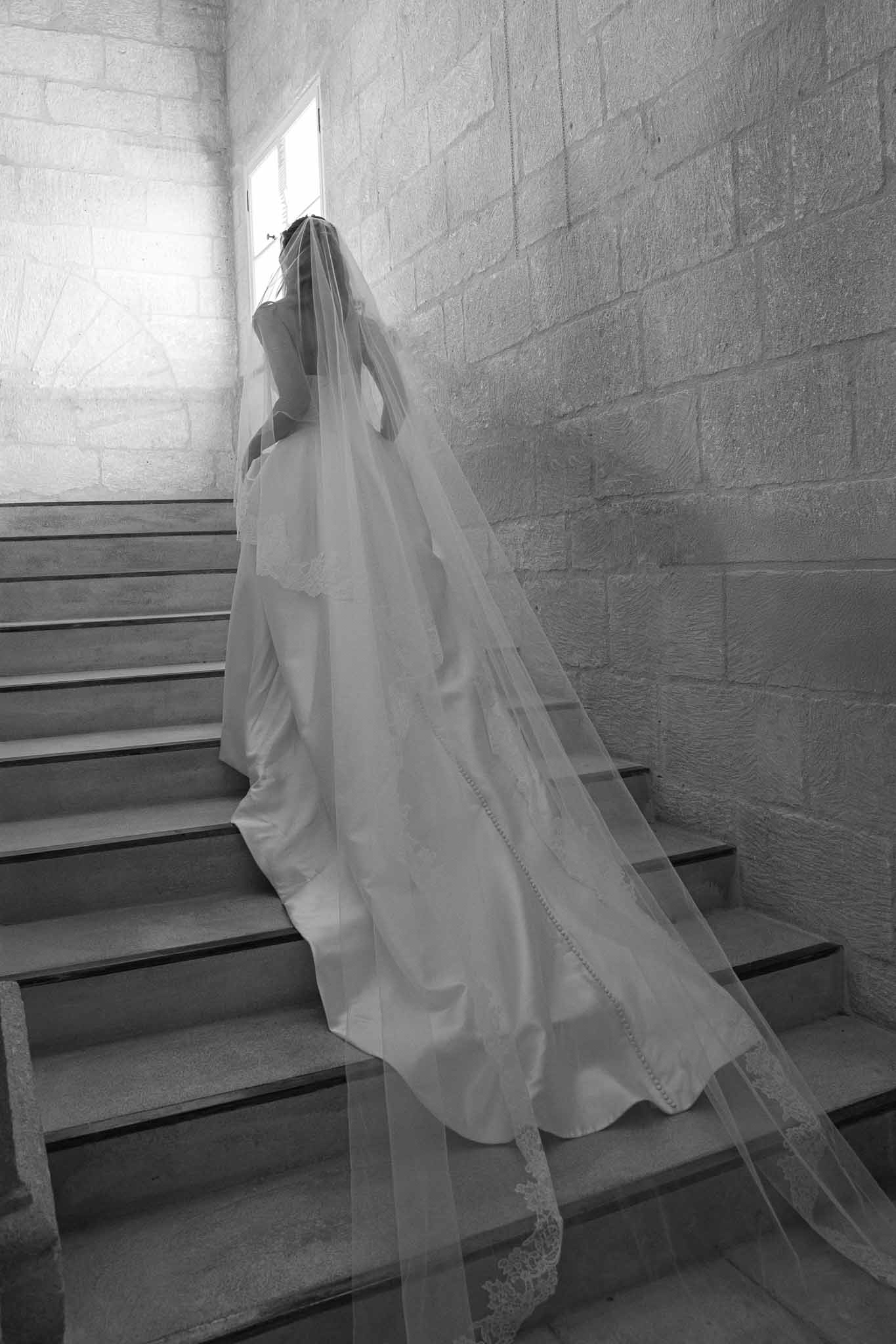 Black and white portrait of bride ascending stone staircase with lace-edged cathedral veil