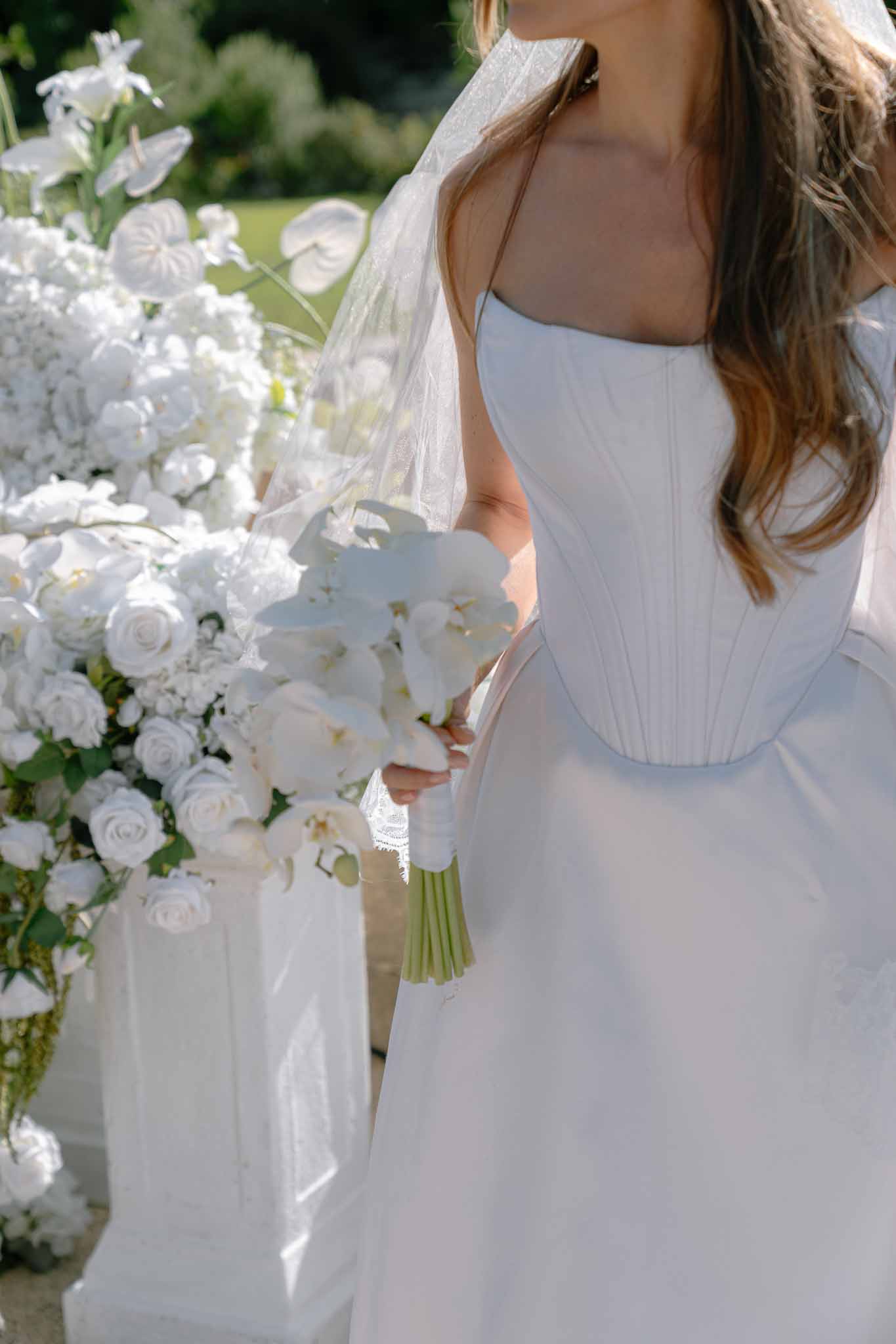 Bride in corset-bodice dress holding white orchid and ranunculus bouquet beside all-white floral installation